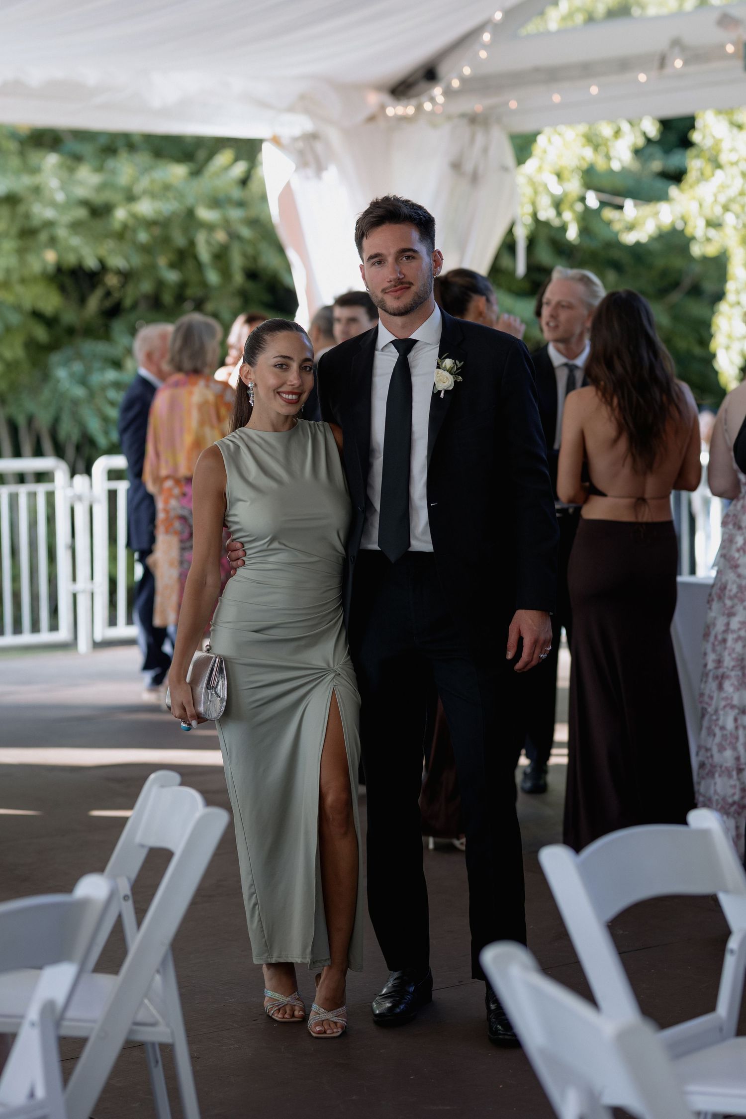 Wedding guests in formal attire socialize under a covered outdoor venue.