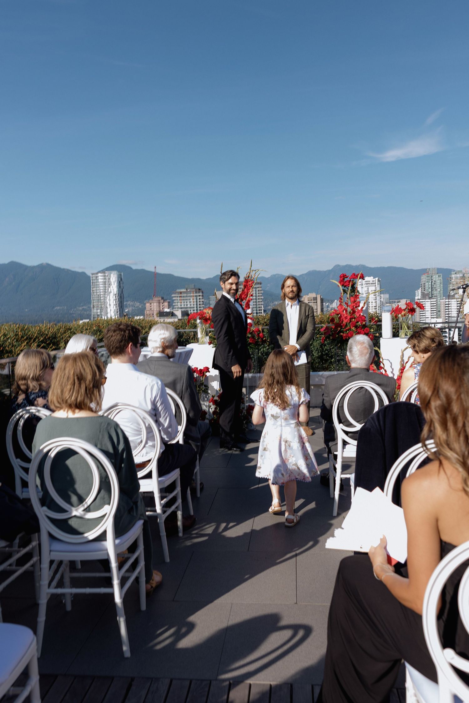 Wedding guests in white chairs gather on a rooftop venue with city skyline views during a sunny ceremony.
