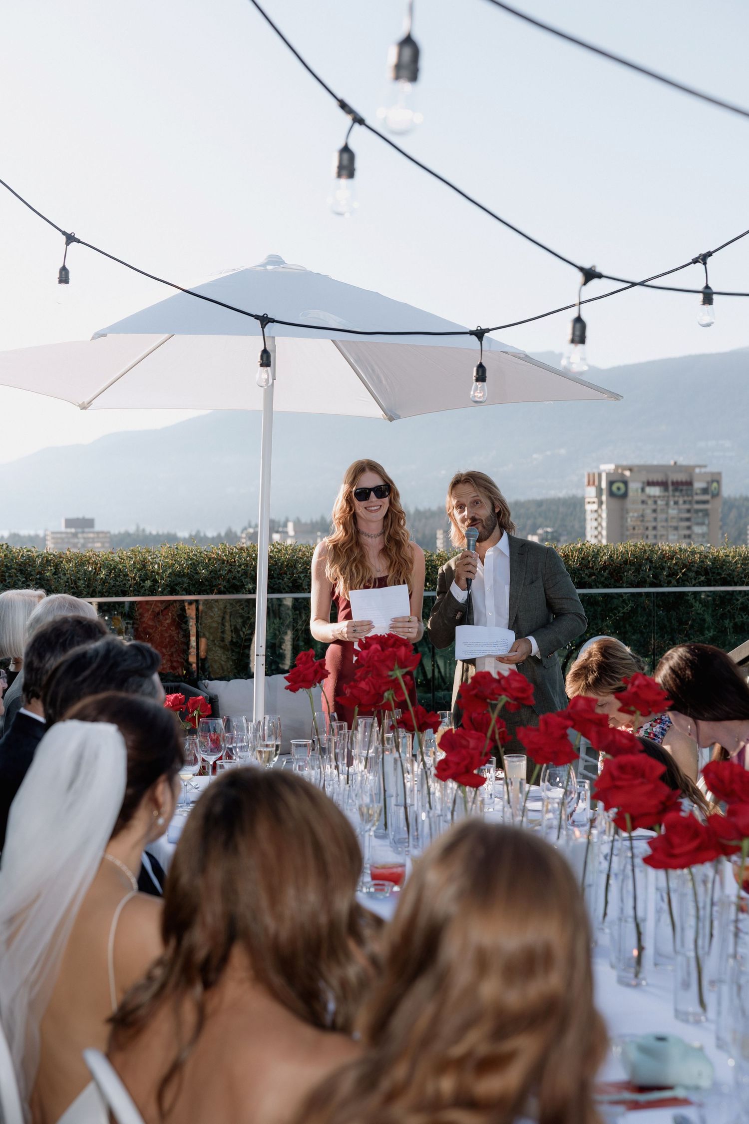 Outdoor wedding reception under string lights with red floral centerpieces and white umbrellas.