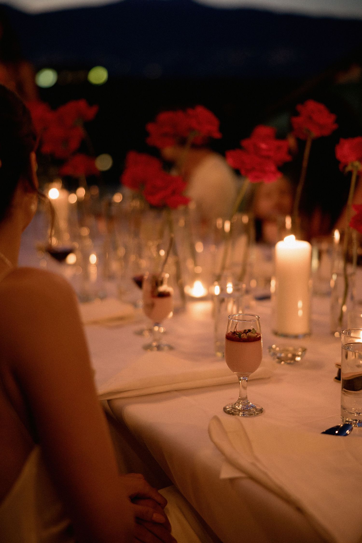 Elegant table setting with white linens, candlelight, and red floral centerpieces at dusk.