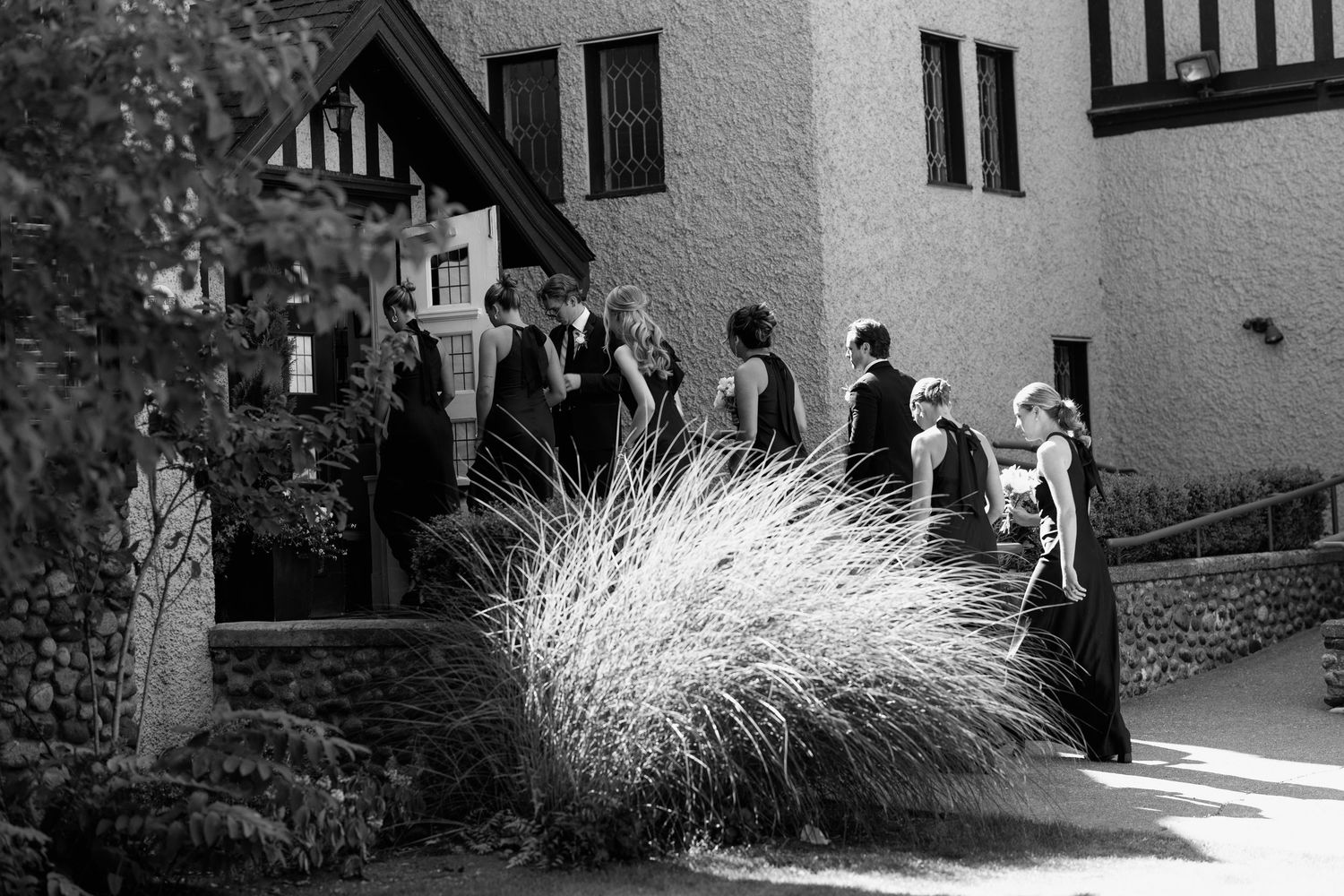 Wedding guests in formal attire gather outside Tudor-style building in black and white.