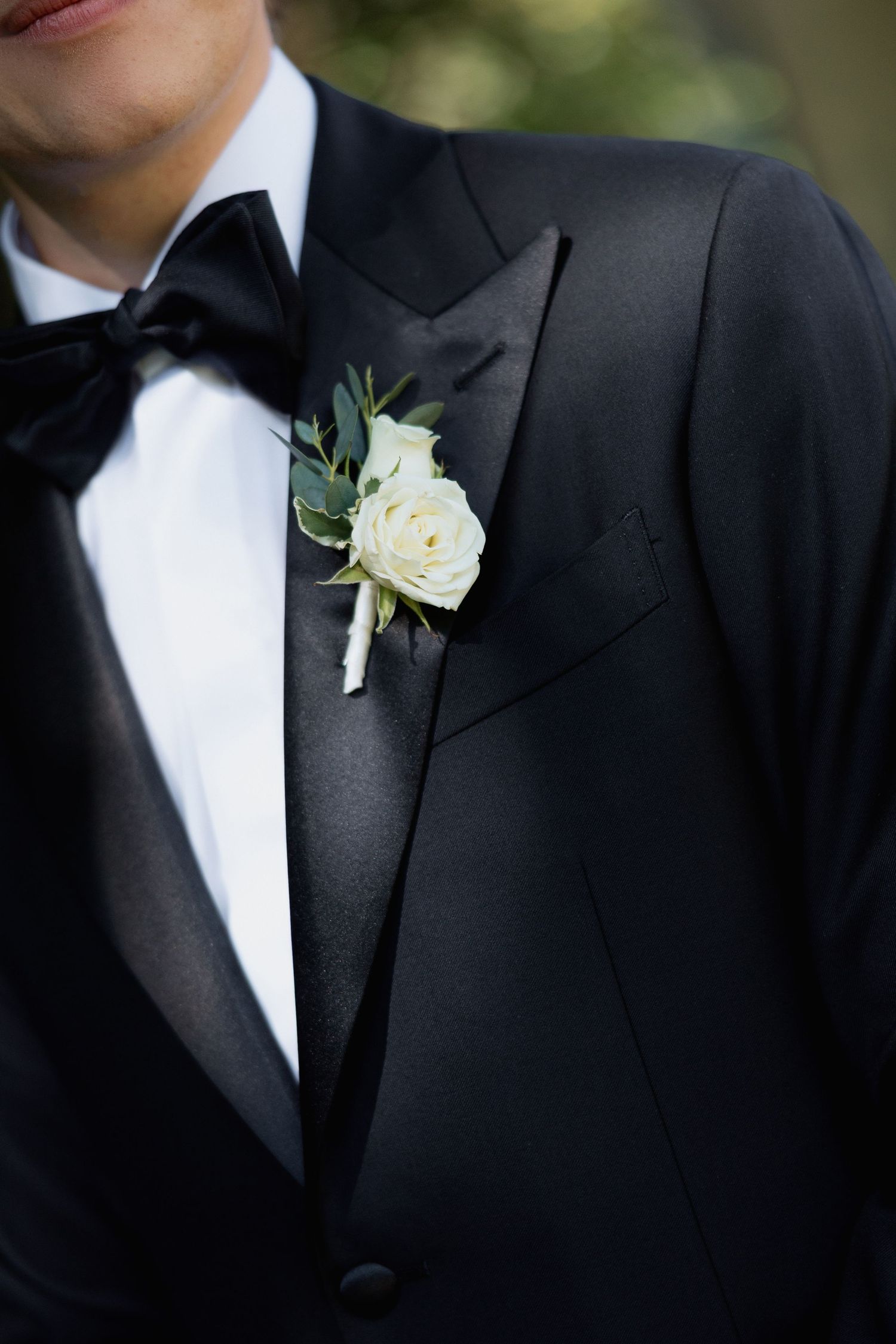 Close-up of groom's formal attire showing white rose boutonniere and black bow tie.