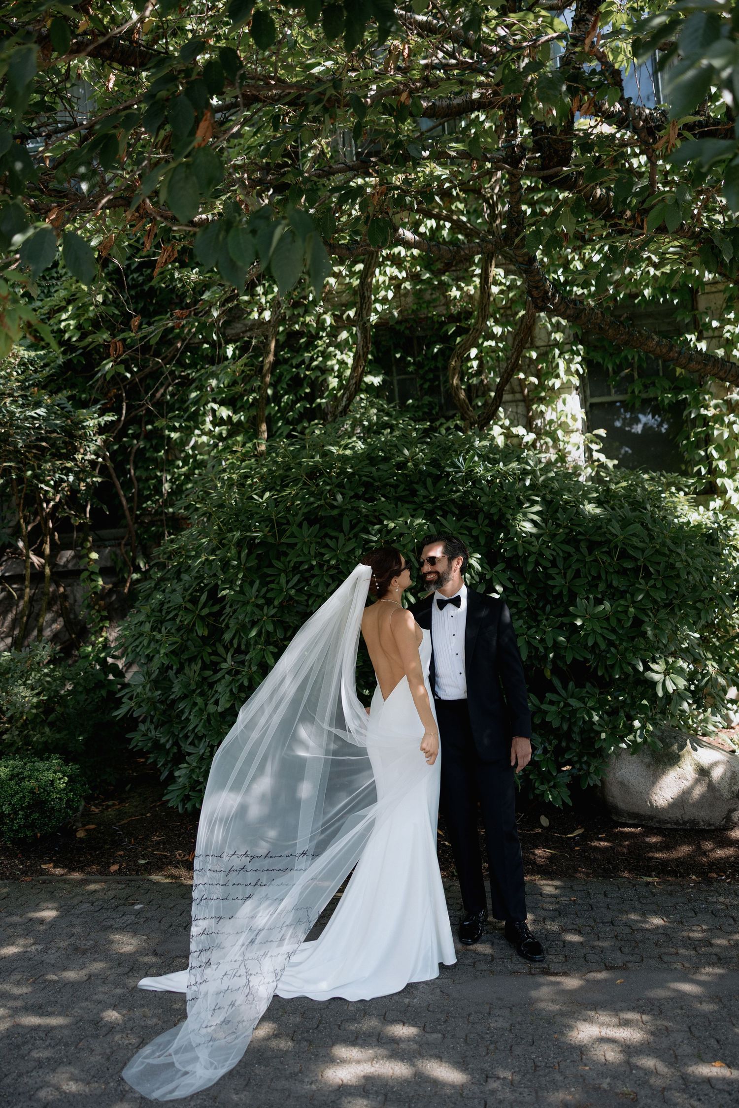 A wedding couple poses under lush green trees with flowing white veil catching the sunlight.