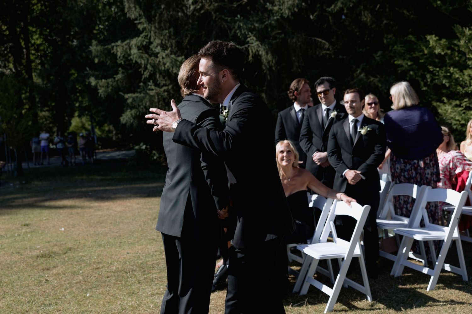 Wedding guests gather outdoors for a ceremony with white chairs arranged on grass under trees.