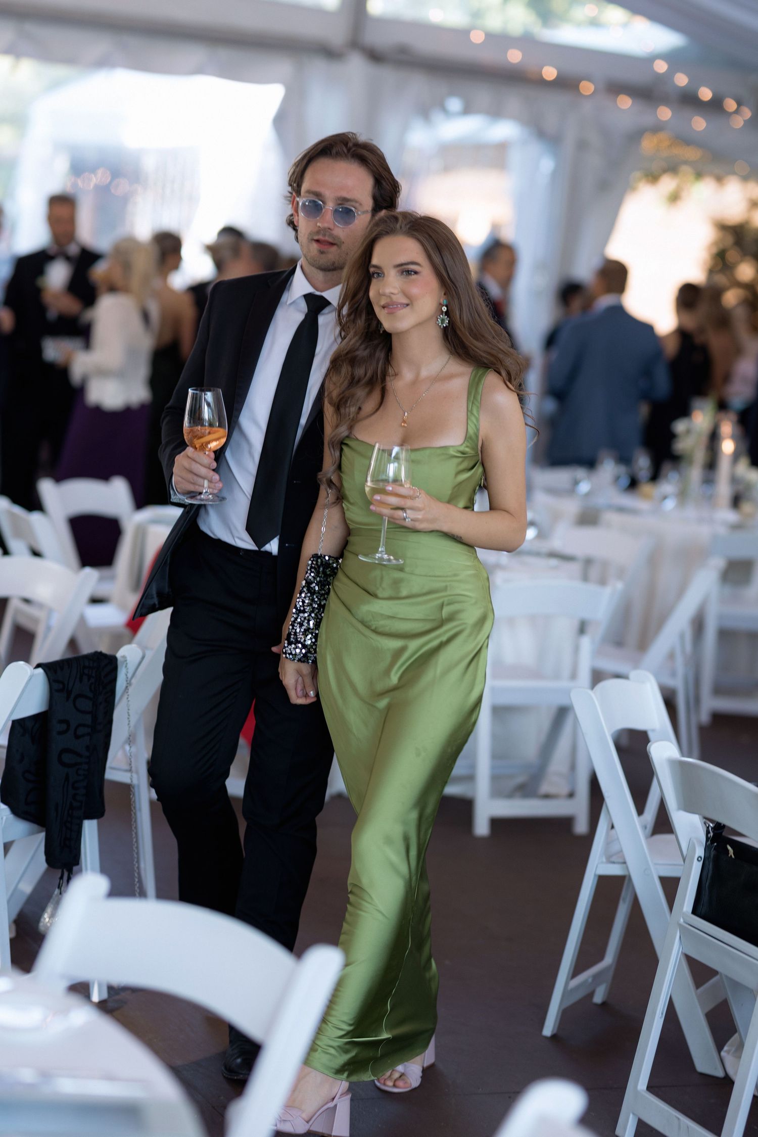 Elegantly dressed couple enjoying drinks at a wedding reception with white folding chairs and string lights.
