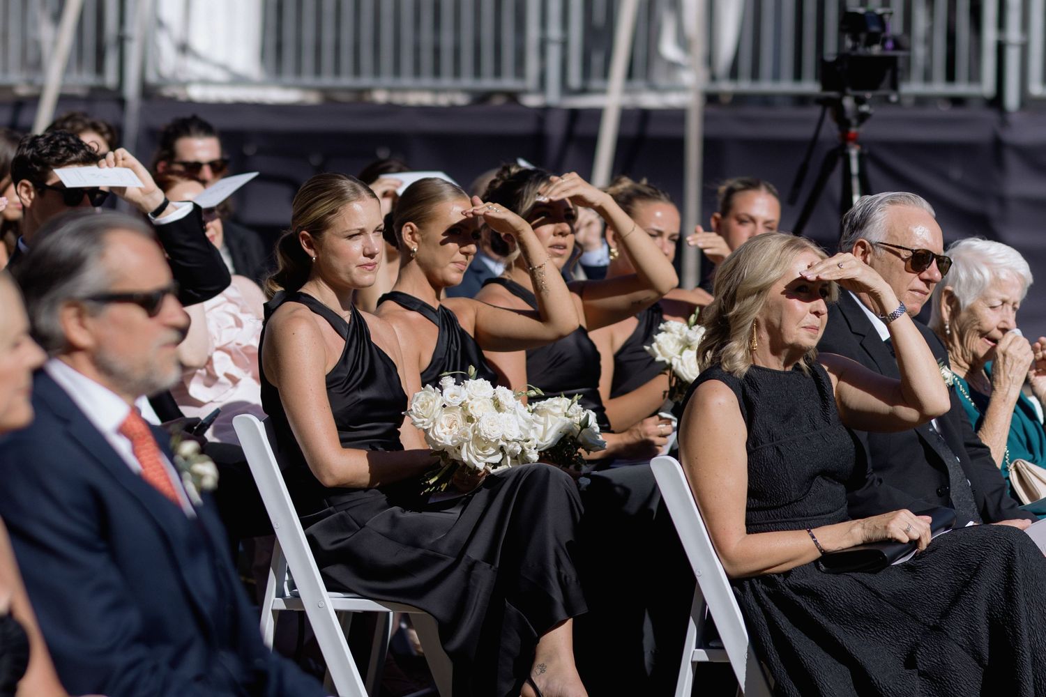 Wedding guests in formal attire watching ceremony from white chairs outdoors.