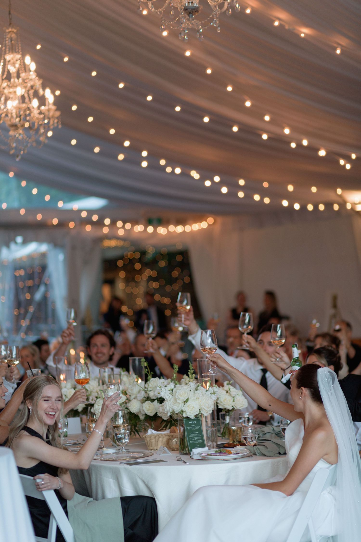 Elegant wedding reception with twinkling lights and white draping overhead as guests raise glasses in celebration.