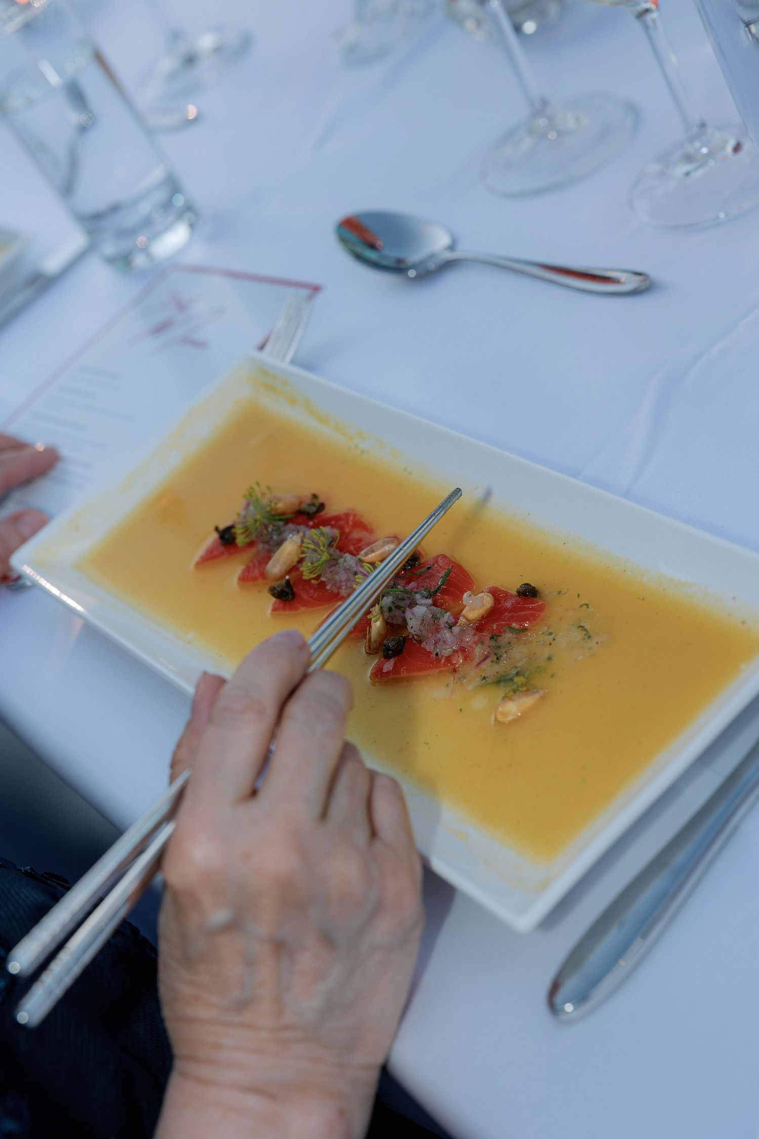 Hand holding chopsticks over yellow soup garnished with red flowers in modern white bowl.