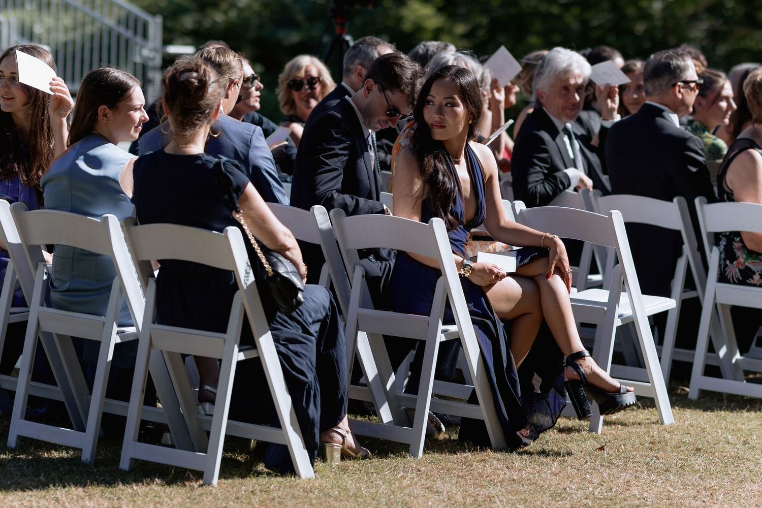 Wedding guests seated in white chairs at an outdoor ceremony on a sunny day.