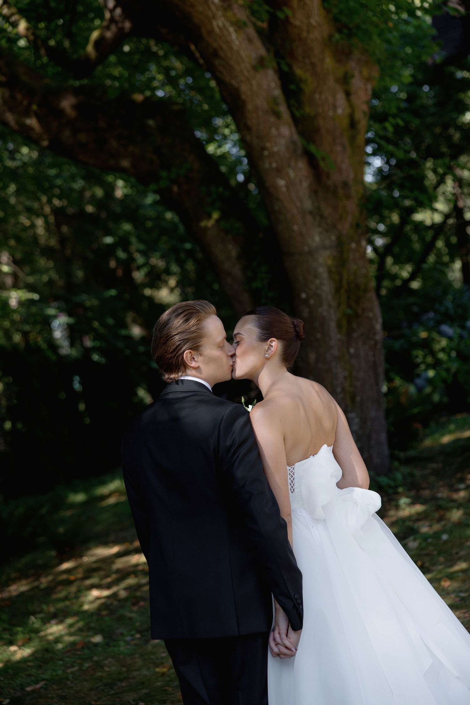 Romantic wedding kiss captured in natural outdoor setting under tall trees.