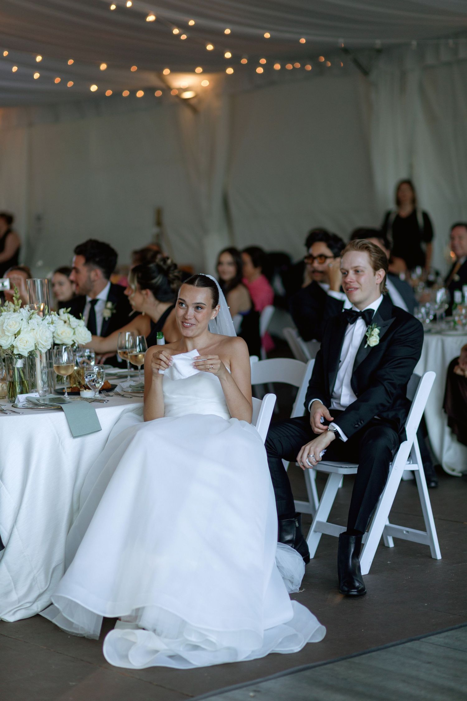 Wedding reception tables set with white linens under string lights in an elegant tent setting.
