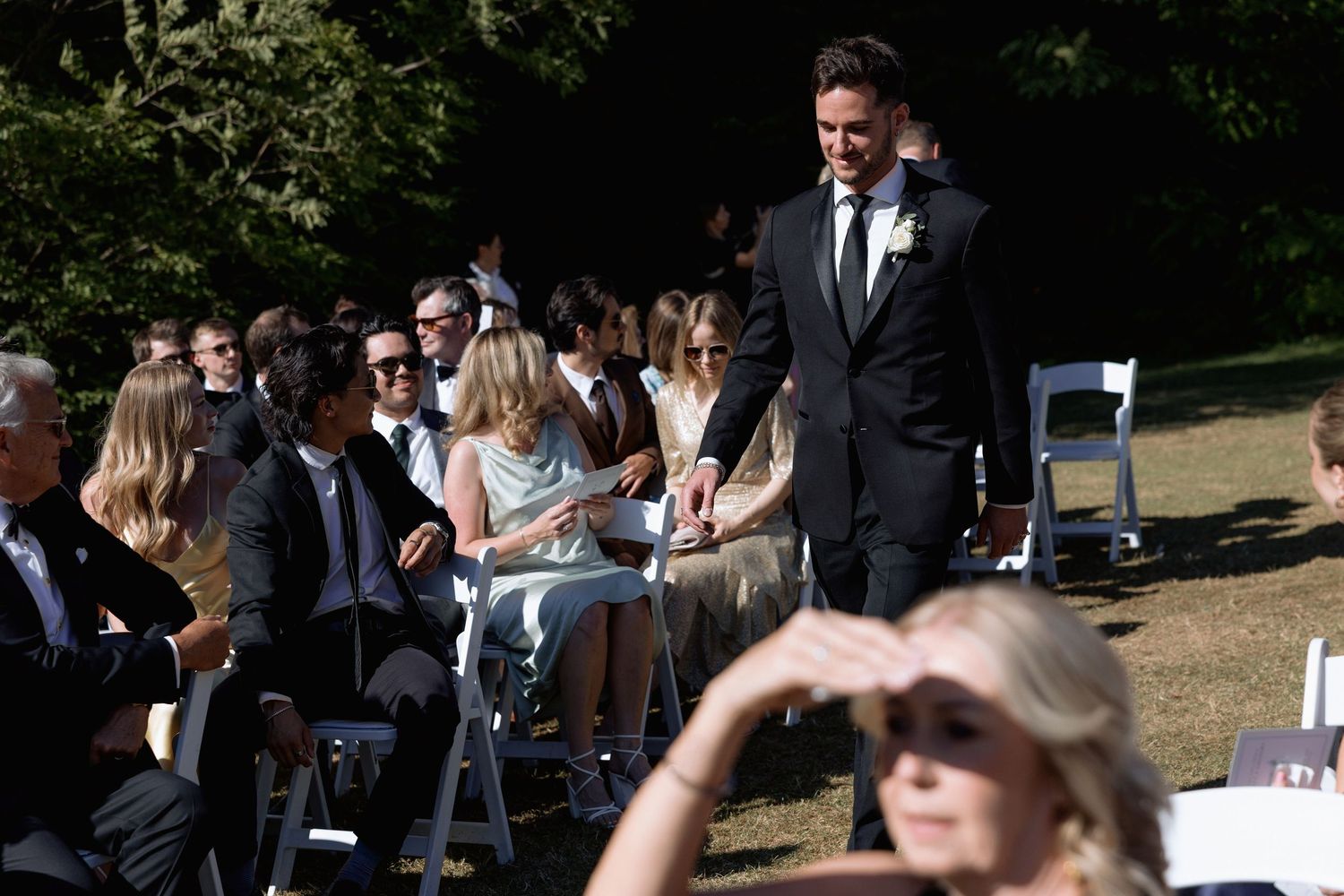 Wedding guests gathering at an outdoor ceremony venue with white chairs and natural surroundings.