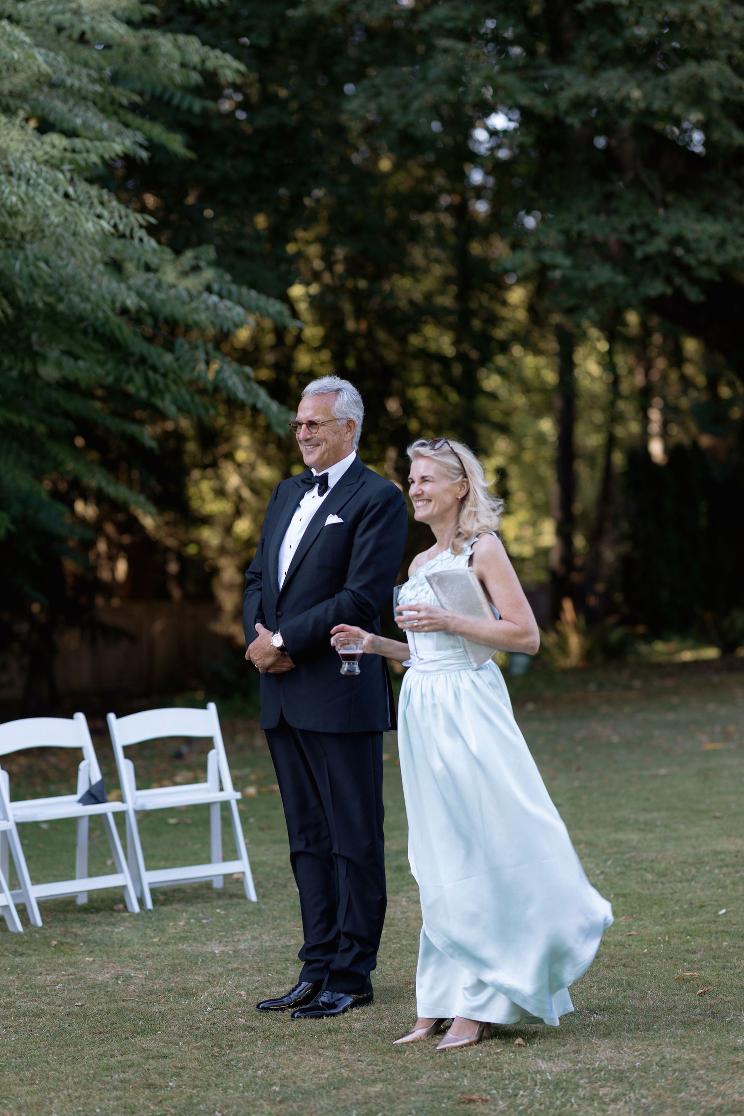 A couple shares a playful moment on grass near white folding chairs.