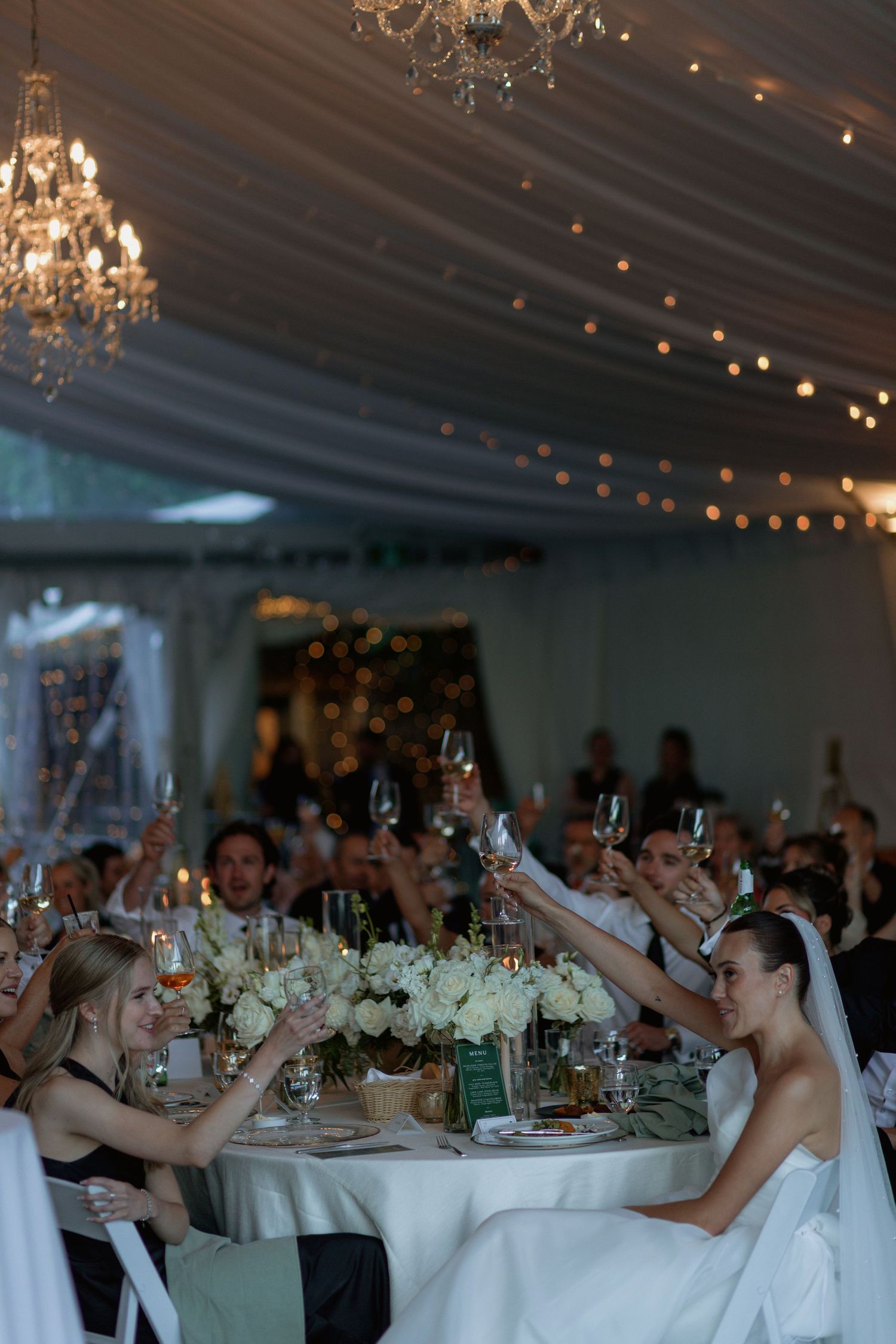 Wedding reception under crystal chandeliers and string lights with guests toasting at decorated tables.
