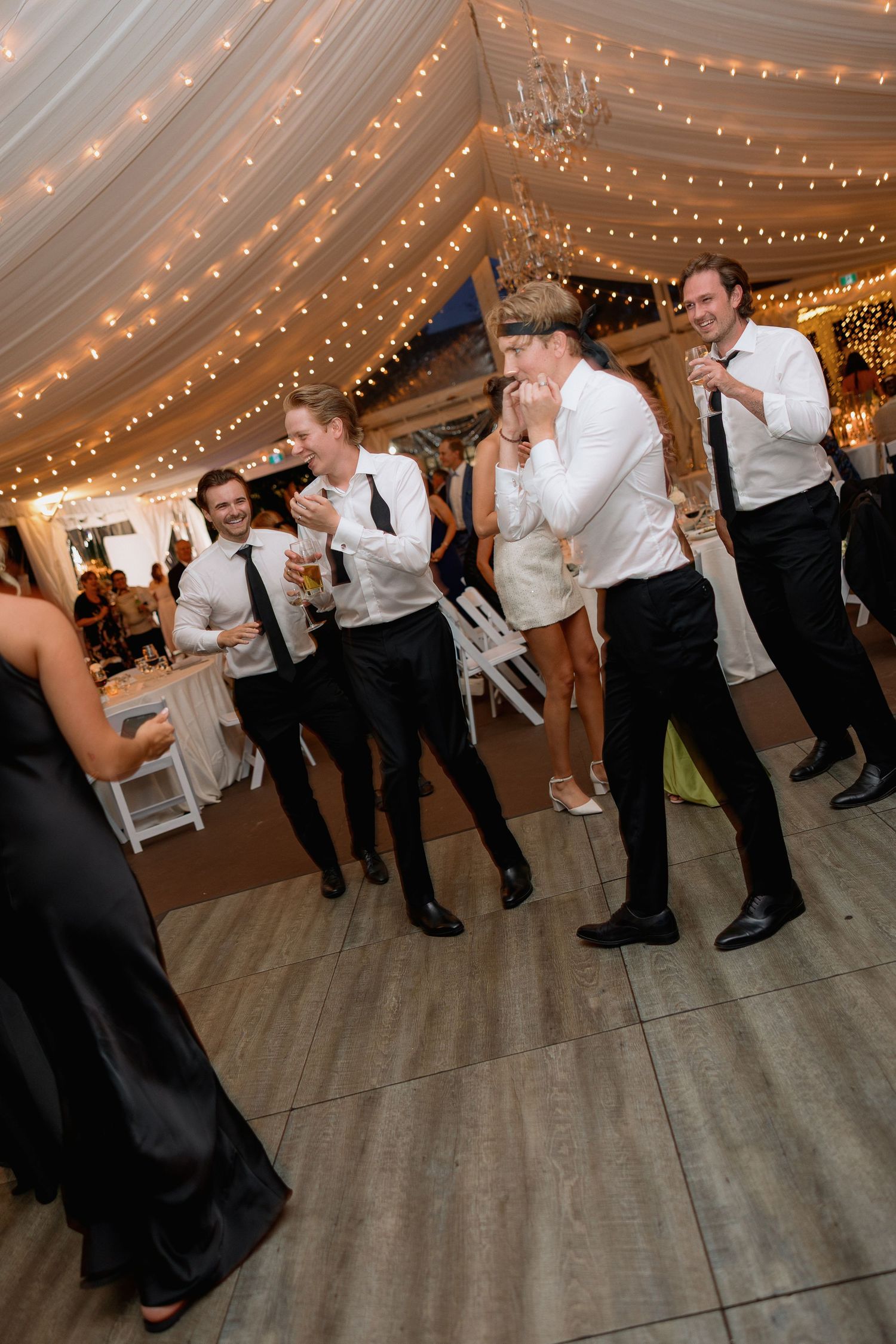 Wedding reception guests dancing in formal attire under string lights in a tent venue.