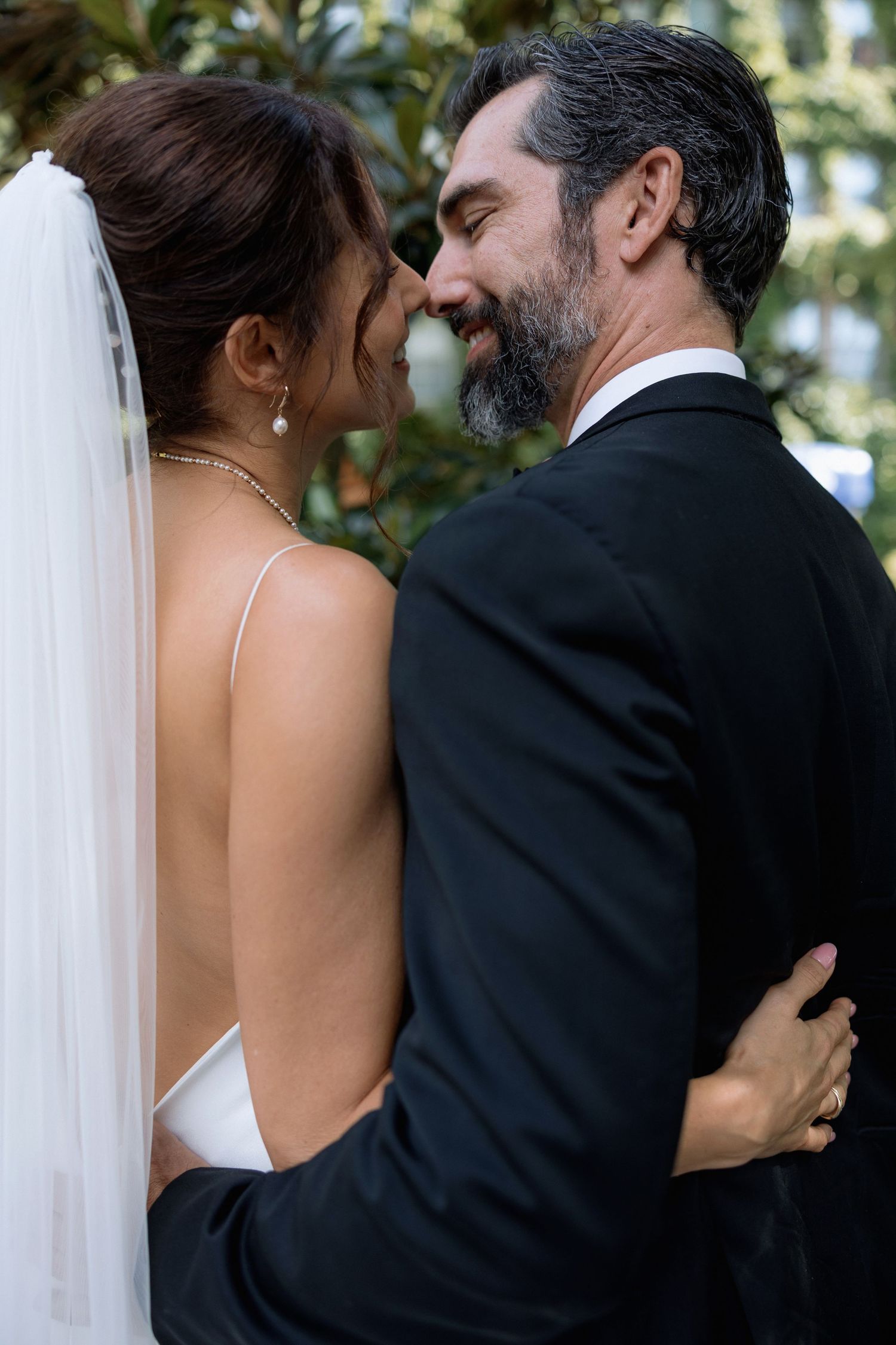 A tender wedding moment captured between newlyweds in formal attire with a white veil against a garden backdrop.