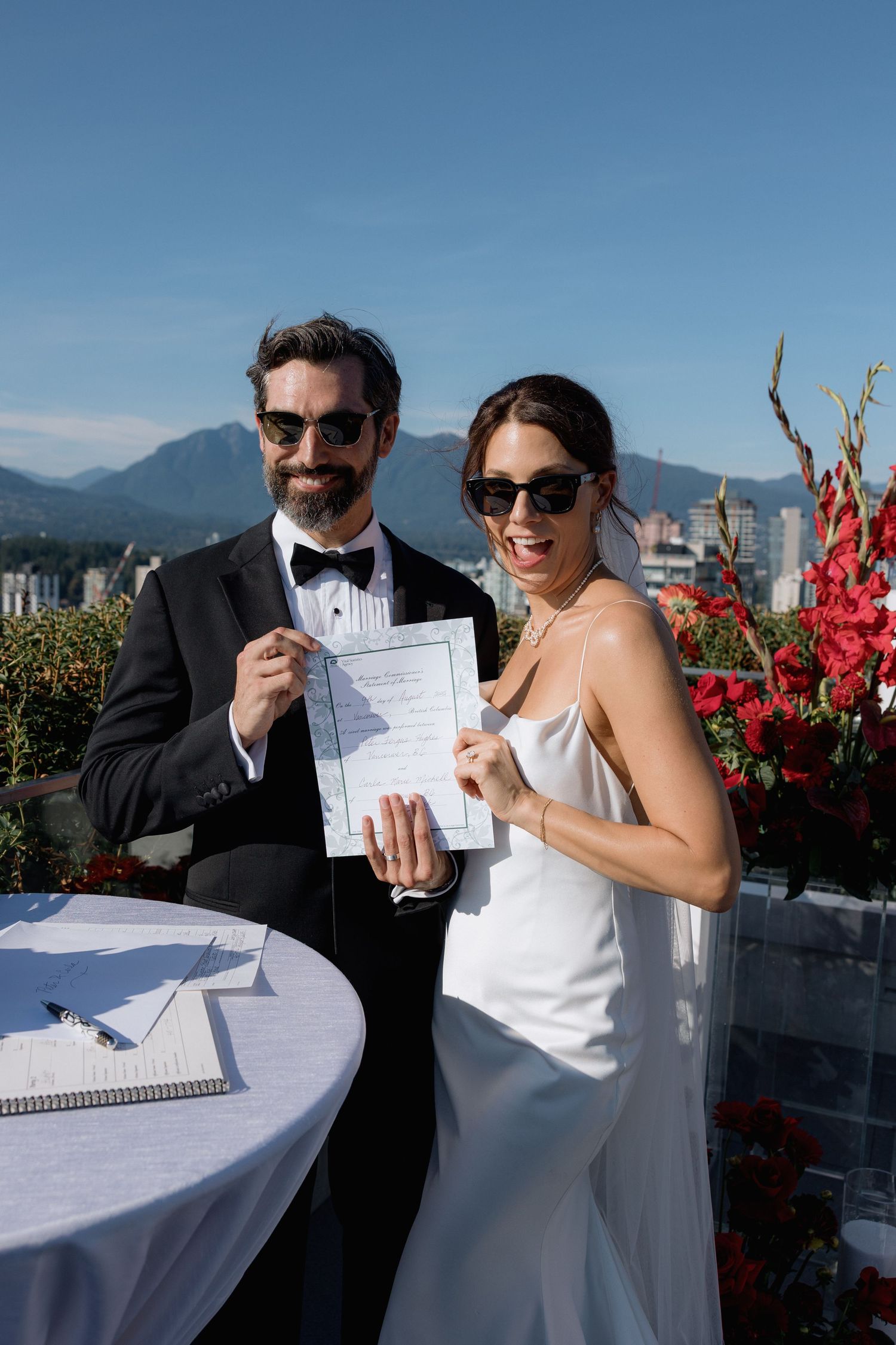 Newlyweds holding marriage certificate on rooftop venue with mountain backdrop.