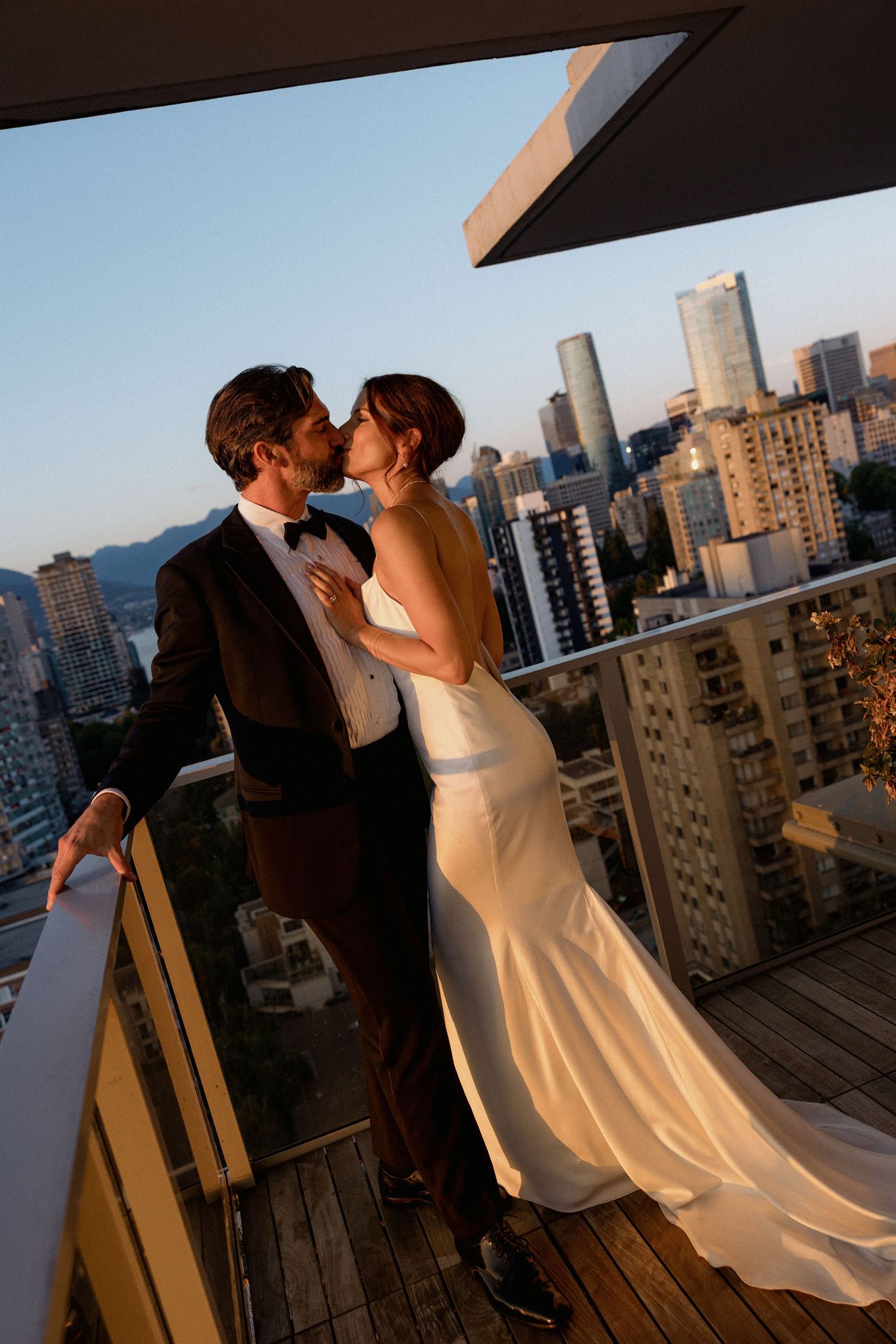 A romantic wedding couple embraces on a rooftop terrace with urban skyline backdrop at sunset.