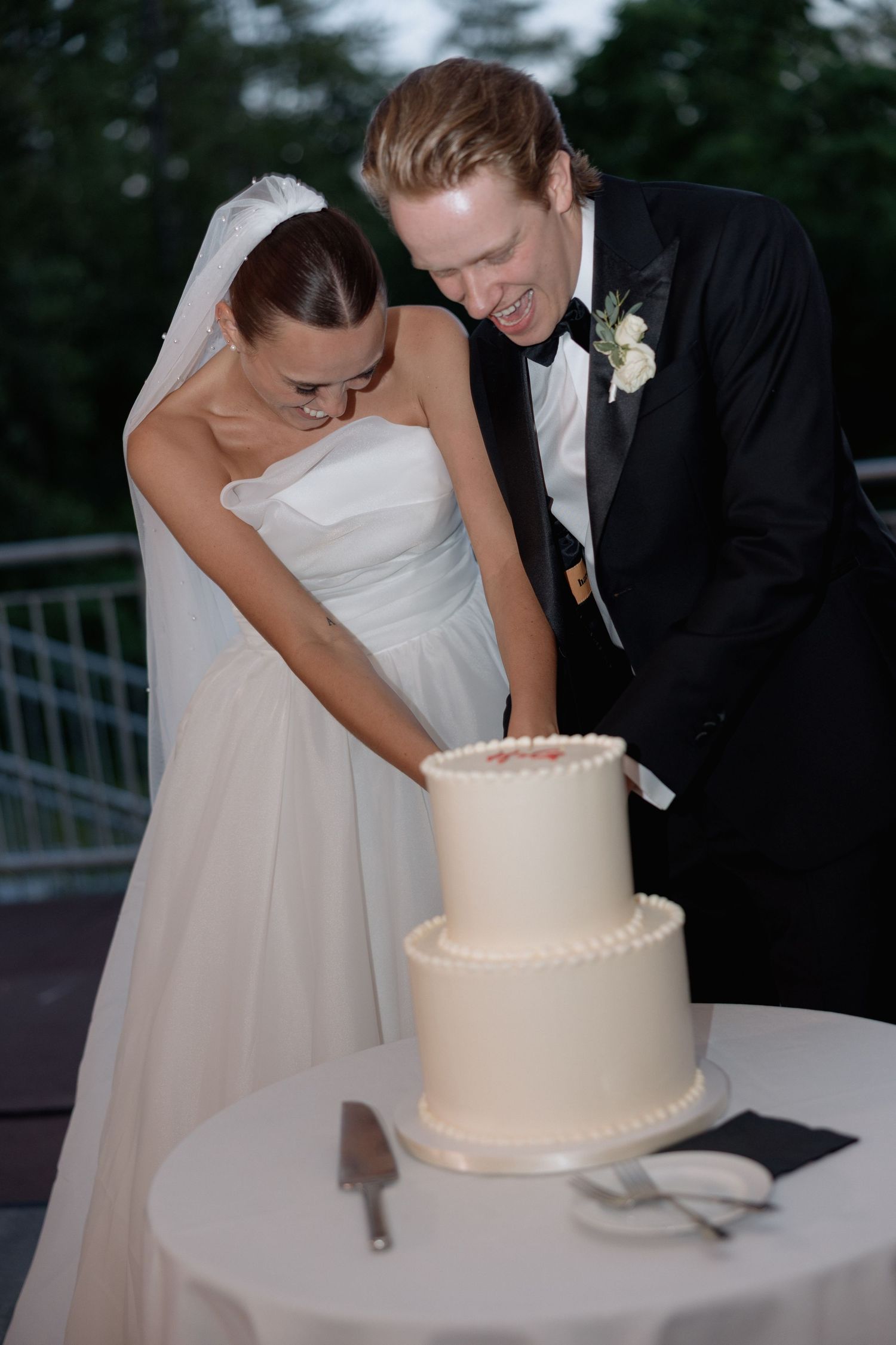 A couple in wedding attire leaning over to look at their wedding cake on a round table.