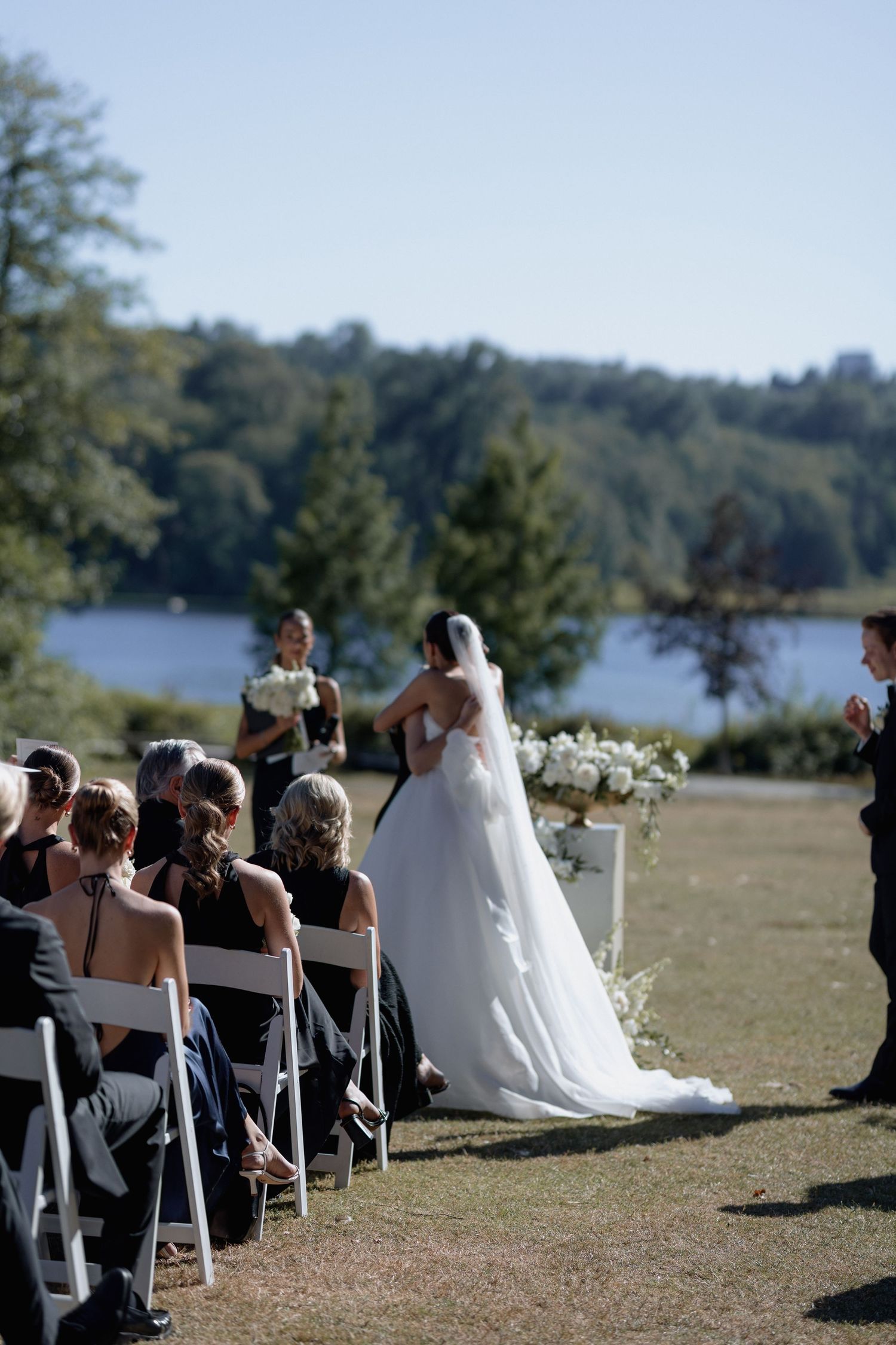 Outdoor wedding ceremony with guests seated on white chairs overlooking a scenic lake and cityscape.