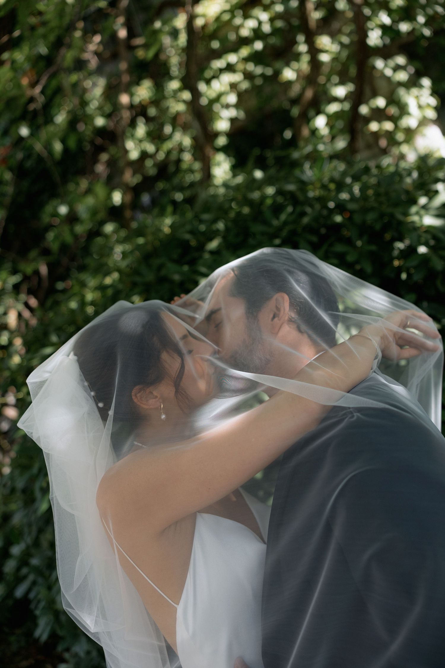 Romantic wedding moment captured under a flowing white veil against green foliage.