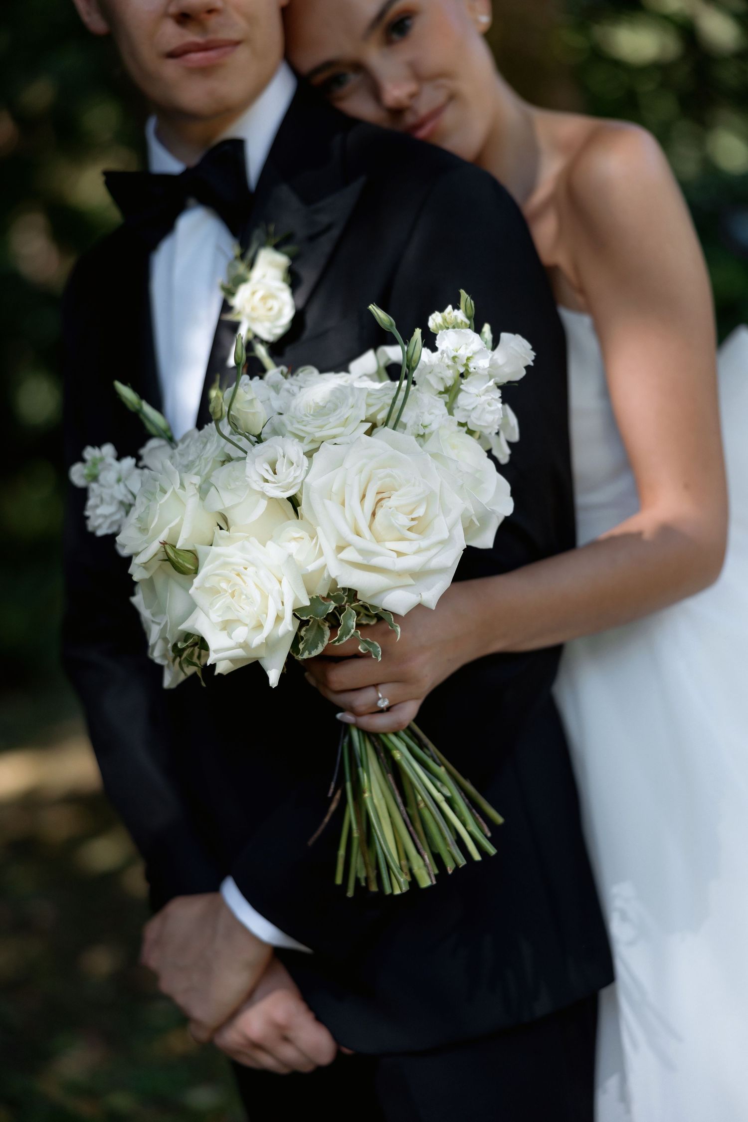 Elegant wedding couple embracing while holding white rose bouquet in wooded setting.