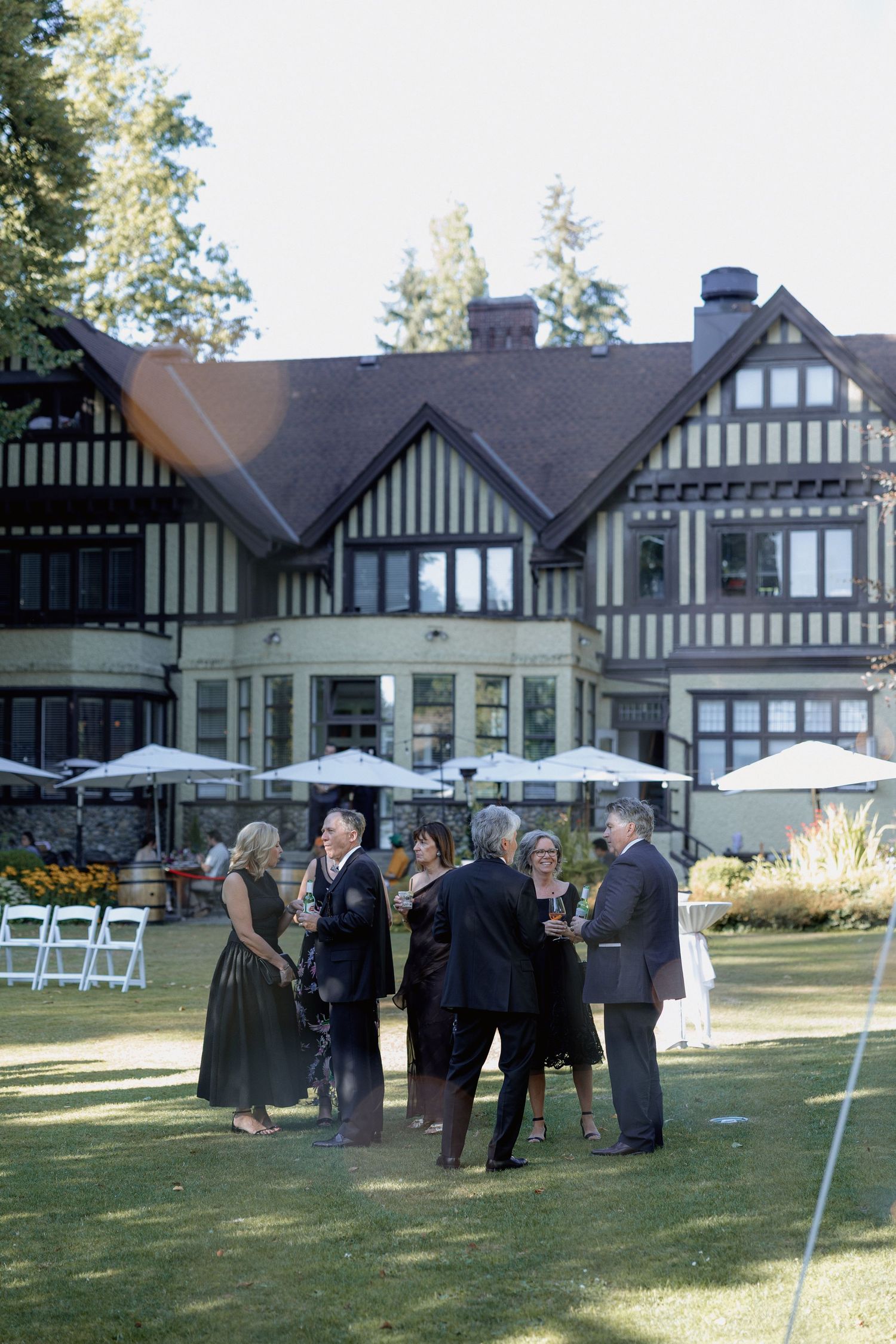 Wedding guests mingling on a lawn with white folding chairs and a Hurt House in Burnaby in the background.
