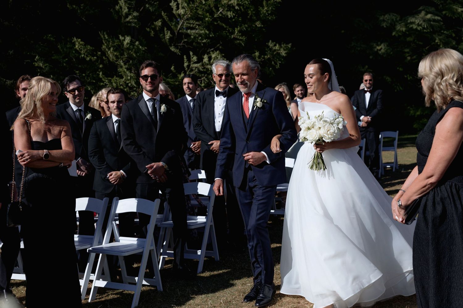 Elegant outdoor wedding ceremony with guests in formal attire and white decorations.