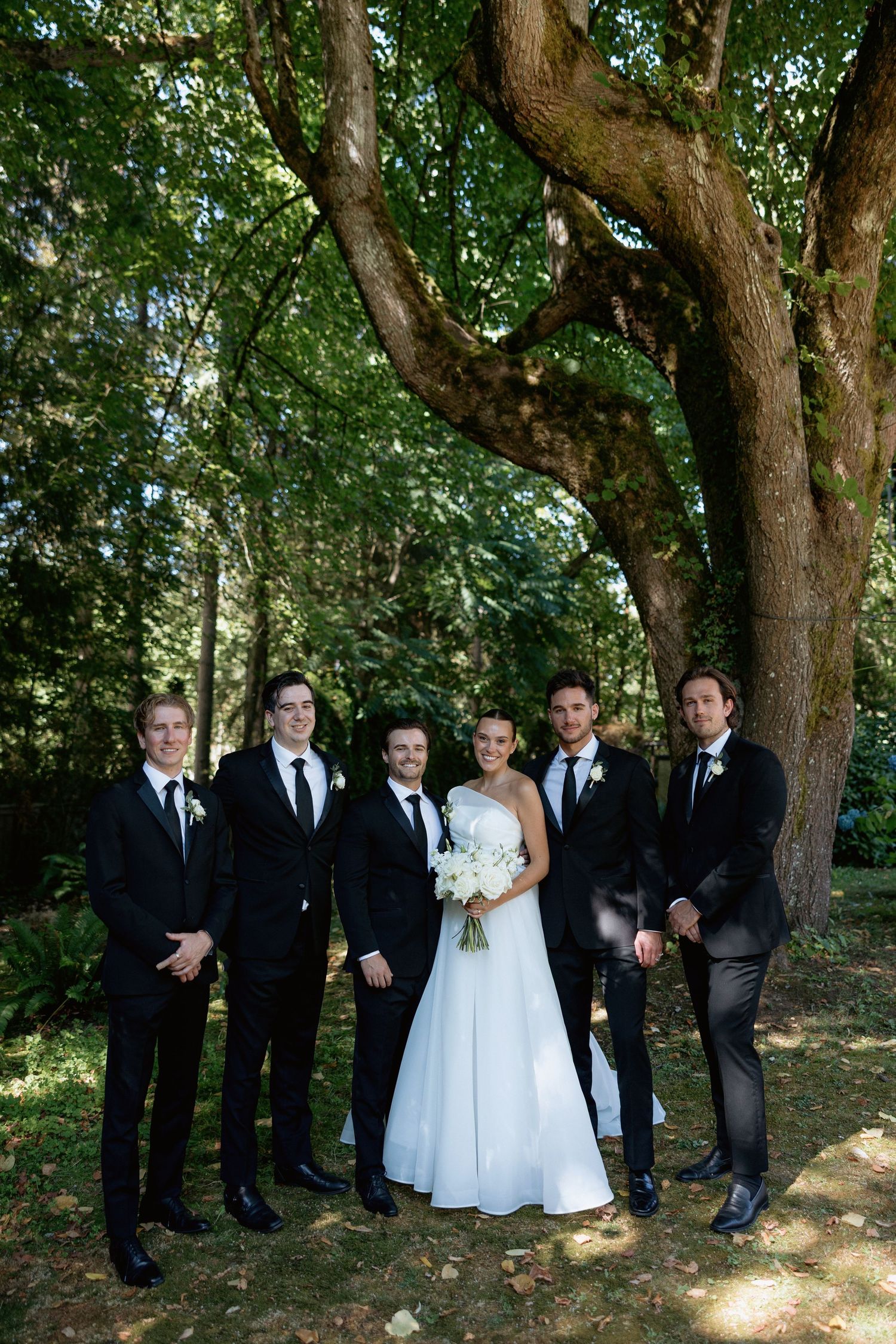 Wedding party in formal attire poses outdoors under a shady tree.