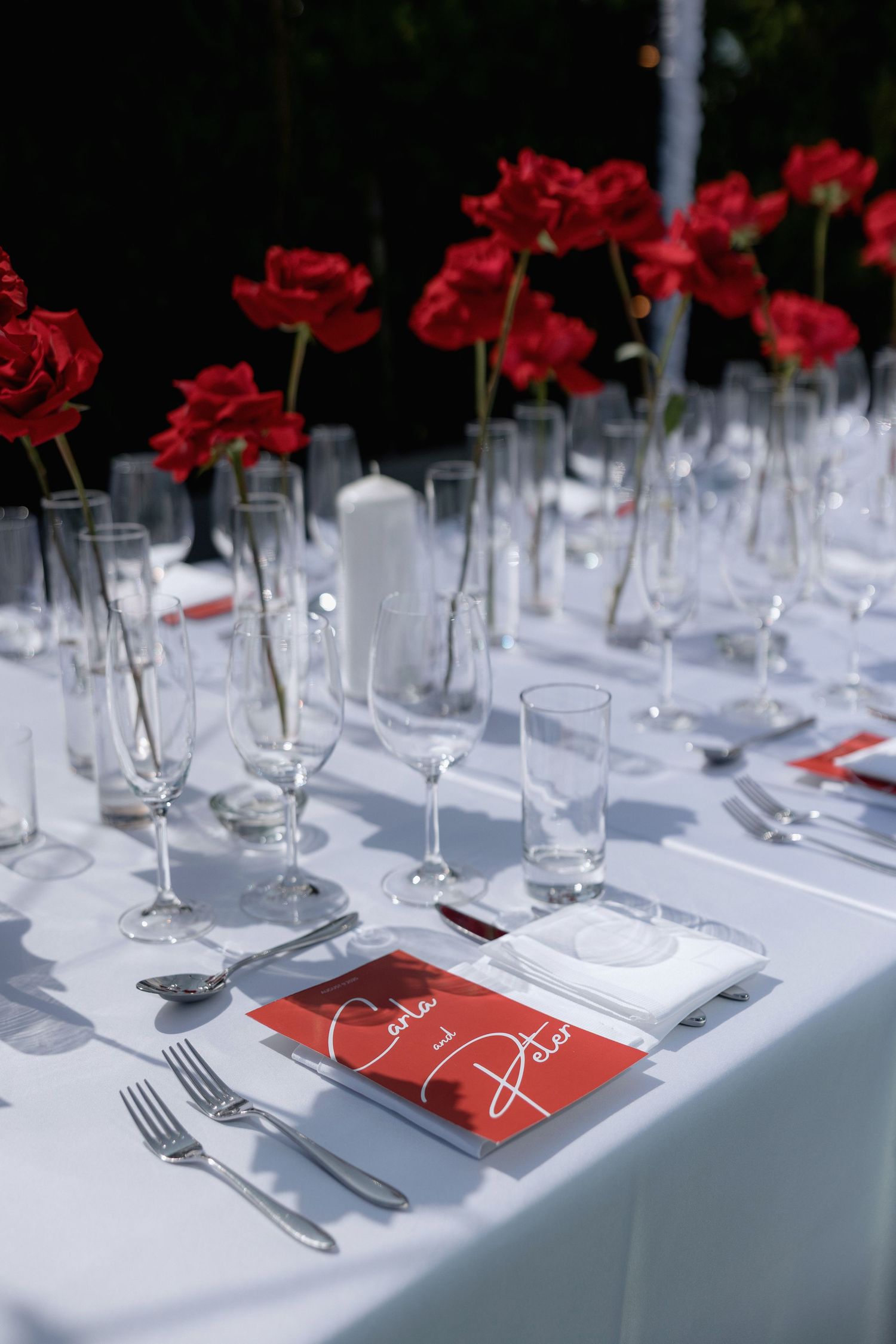 A formal table setting with red roses in vases, white tablecloth, and wine glasses arranged in a row.