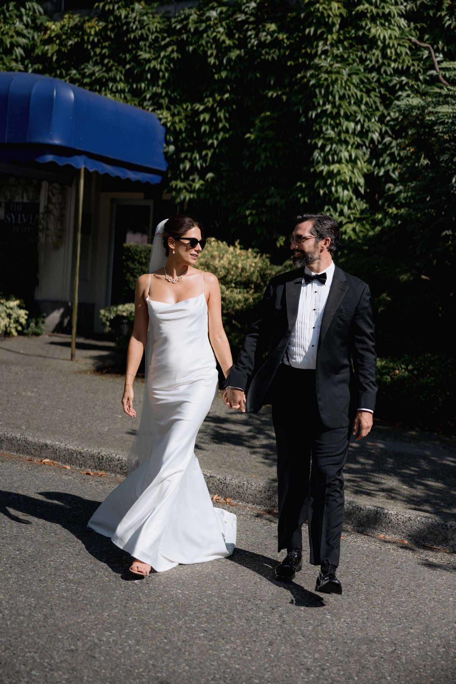 A bride in white dress and groom in tuxedo walk together on a paved outdoor path in front of the Sylvia Hotel in Vancouver
