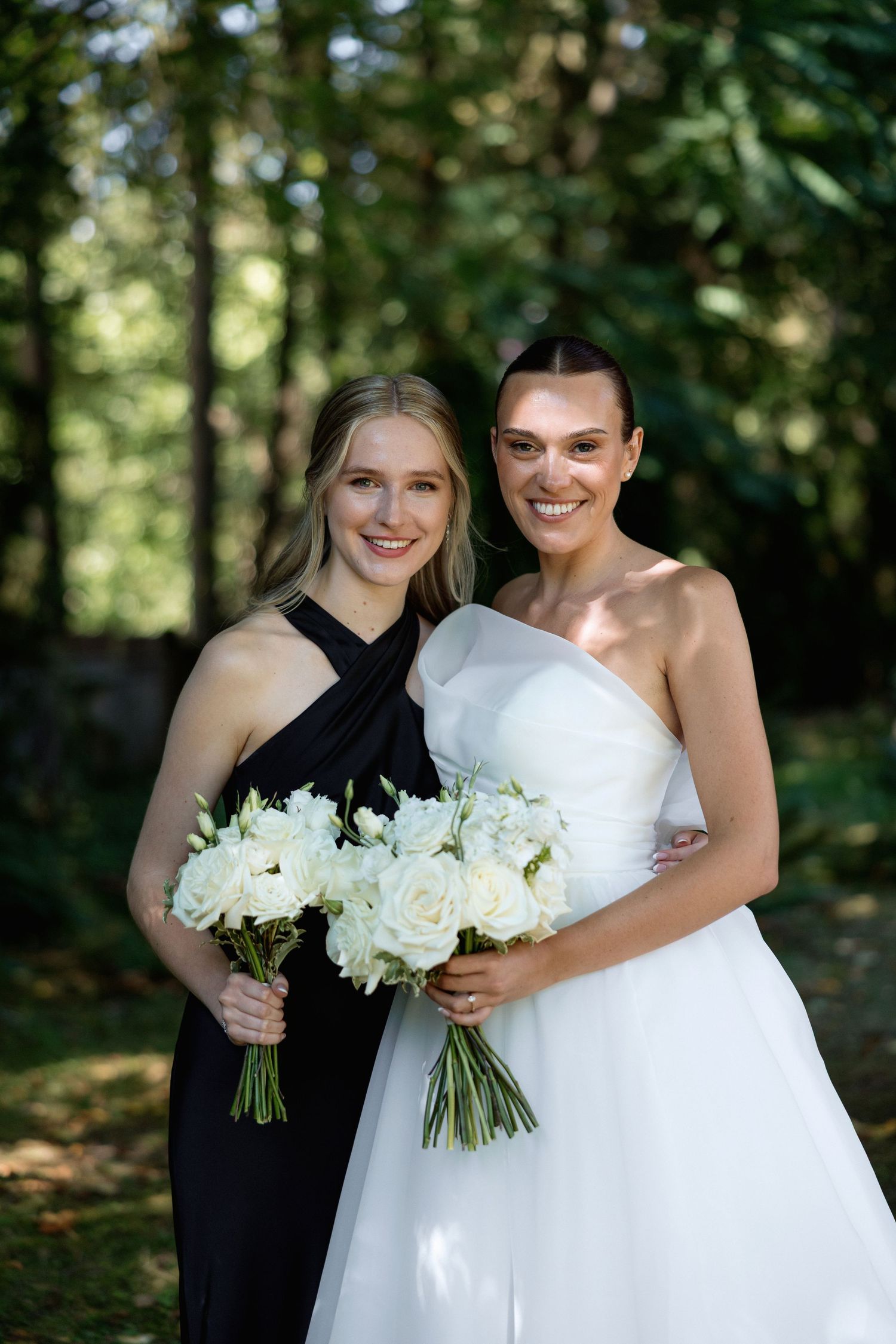 Wedding party members smile while holding white rose bouquets in a wooded outdoor setting.