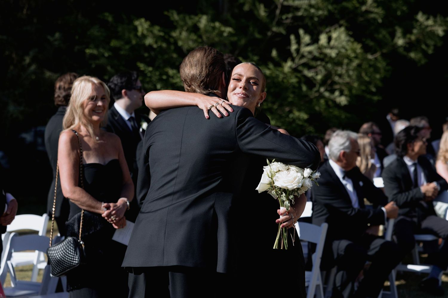 Wedding guests gather outdoors for a ceremony with white chairs arranged on a grassy lawn.