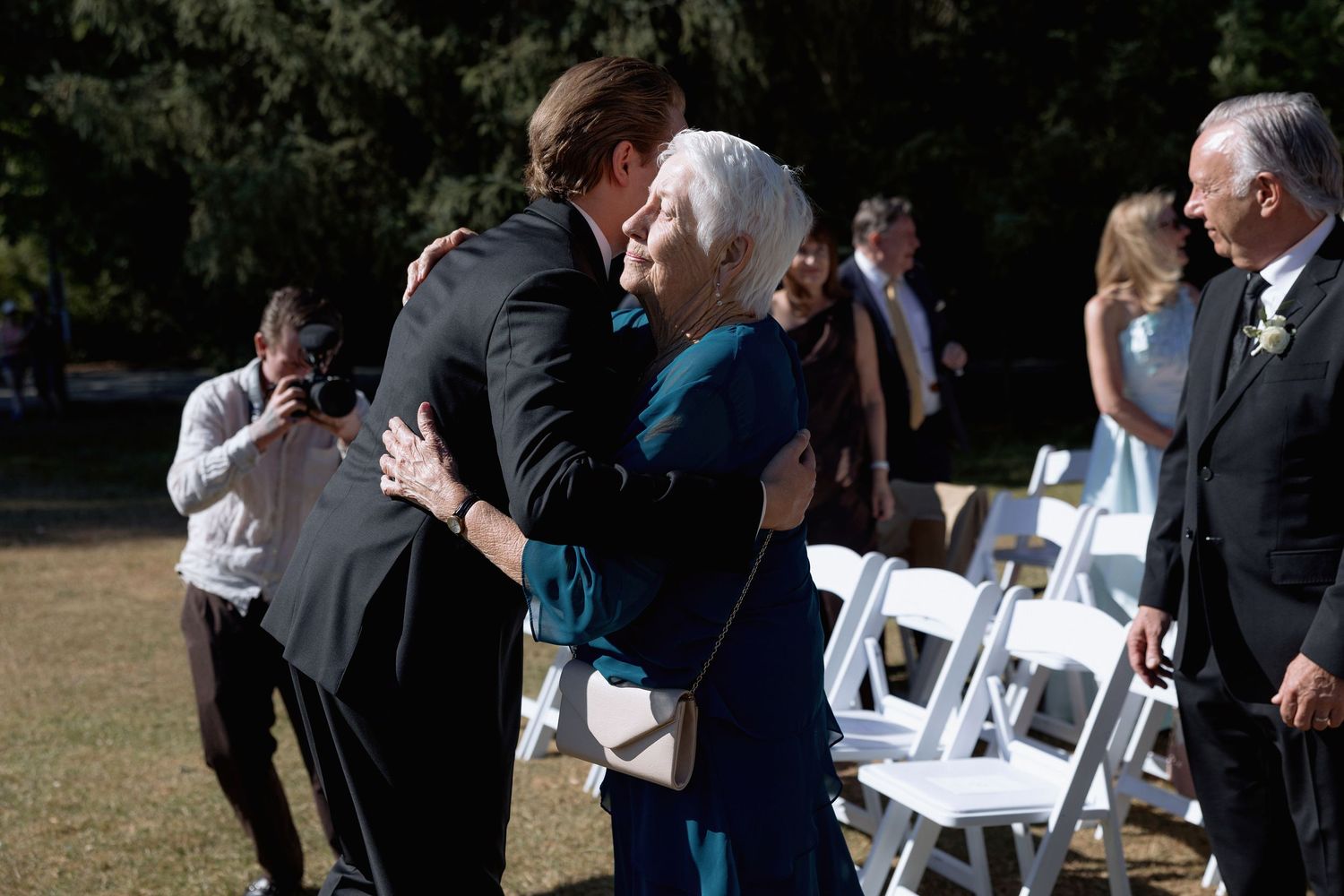 Wedding guests interact near white folding chairs set up on a grass lawn.
