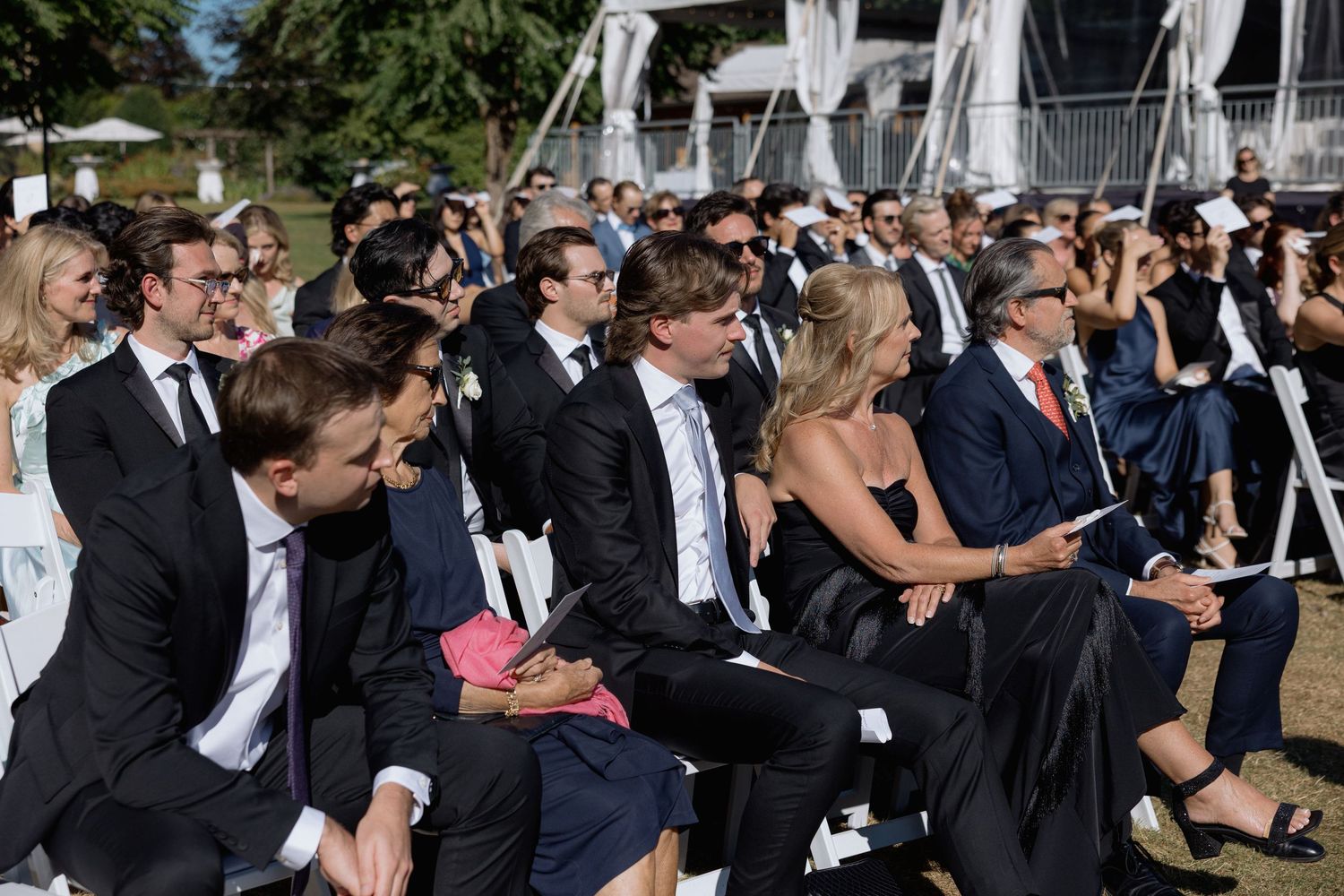 Wedding guests in formal attire sit in rows of white chairs during an outdoor ceremony.