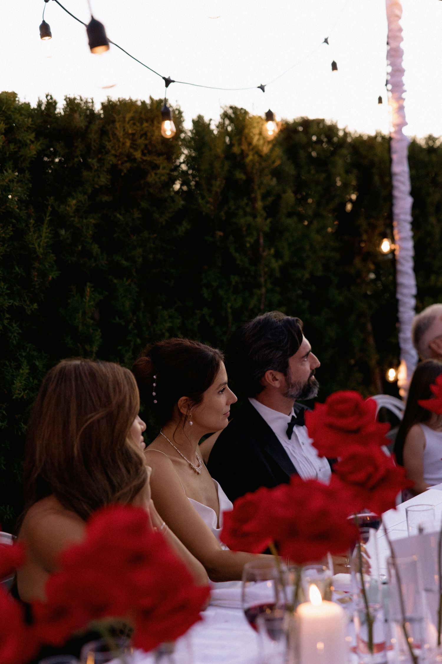 Outdoor wedding reception table decorated with red roses and string lights at sunset.