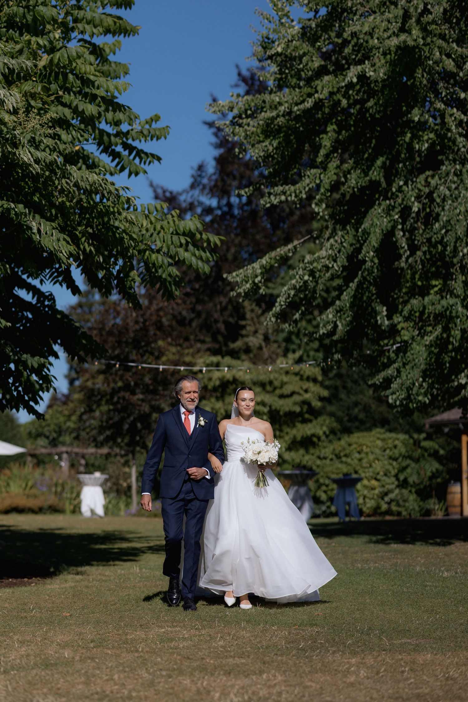 A wedding party walks across a sunny lawn with evergreen trees in the background.
