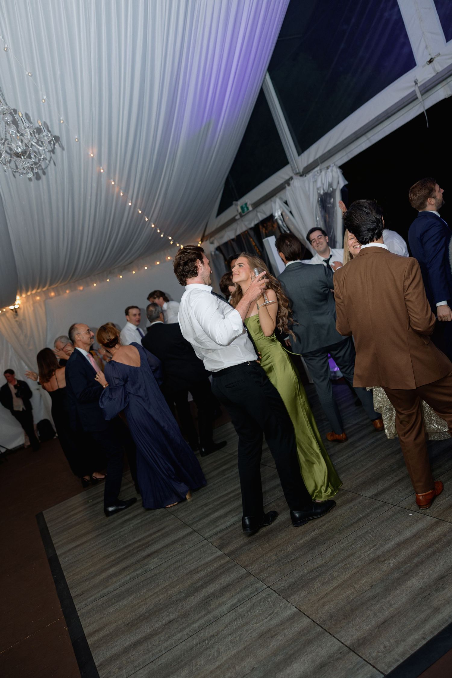 Wedding guests dancing under a tent with purple uplighting and string lights.