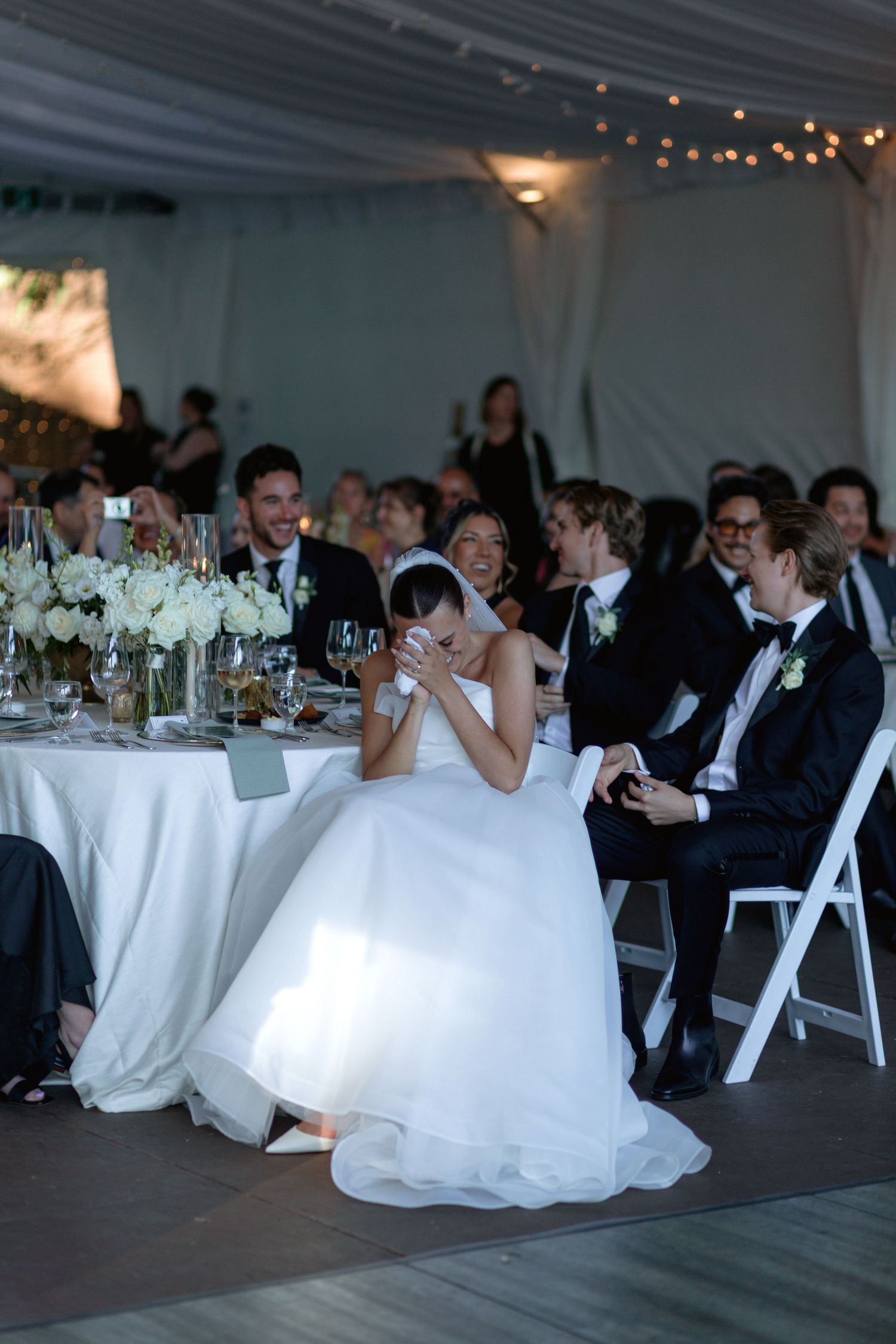 Wedding guests seated at elegantly decorated tables in a white tent venue with string lights overhead.
