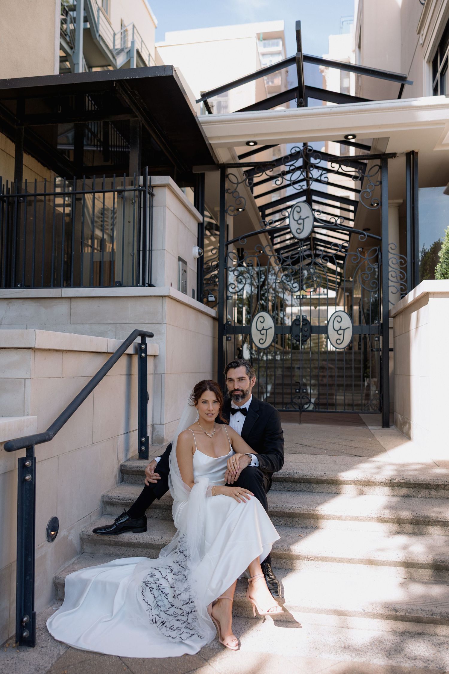 A wedding couple sits together on elegant white stone steps with decorative metal railings.