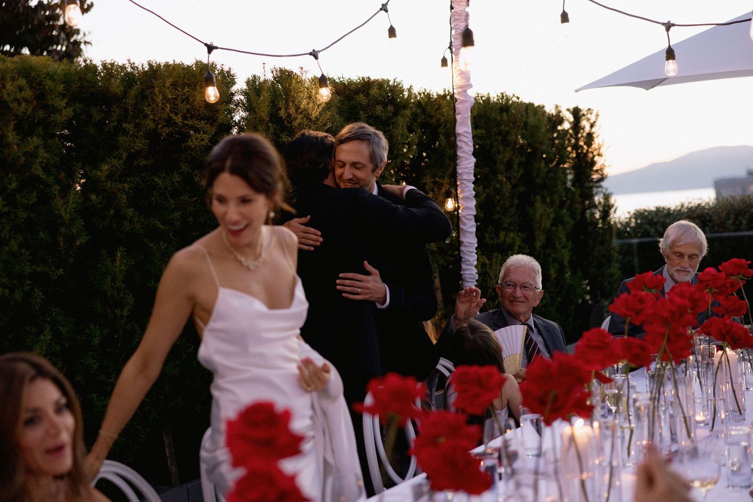 Wedding reception setup with string lights and red floral decorations at dusk.