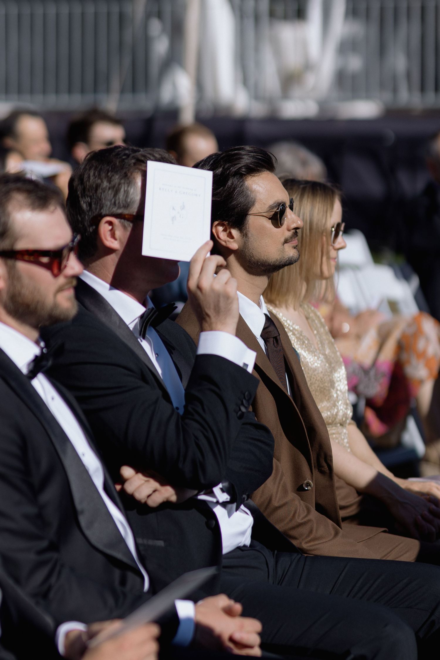 Formally dressed wedding guests holding up programs during outdoor ceremony.