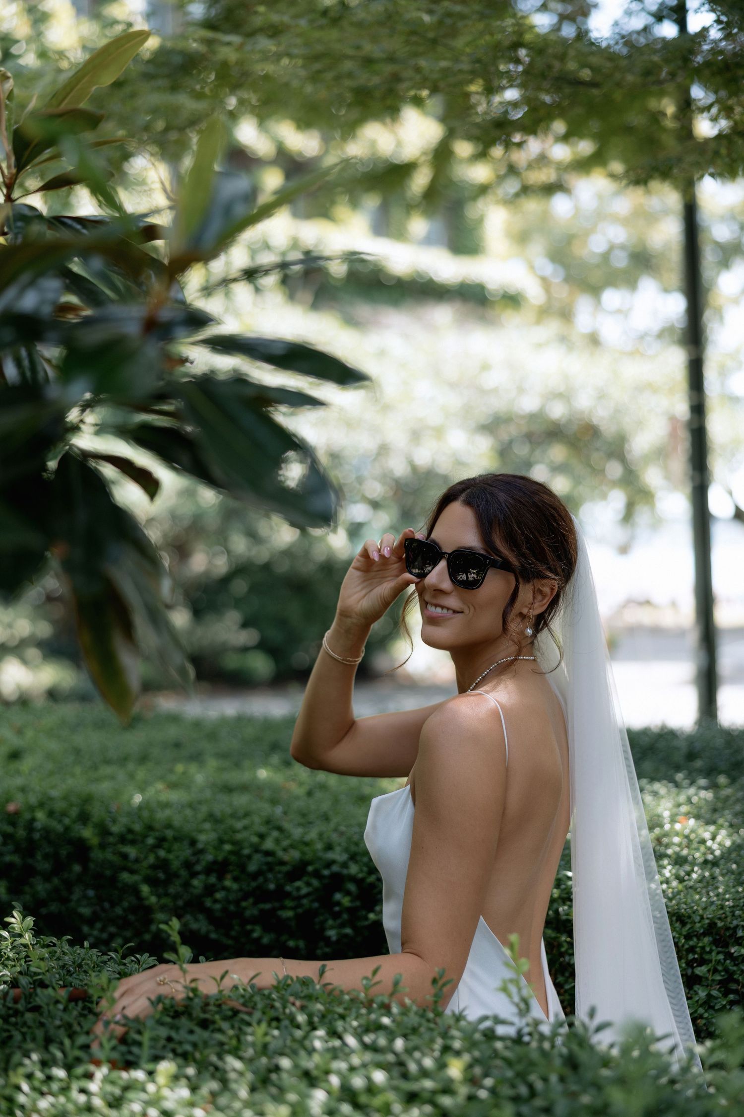 A bride in a white backless dress and sunglasses poses in a garden setting with greenery and natural light.