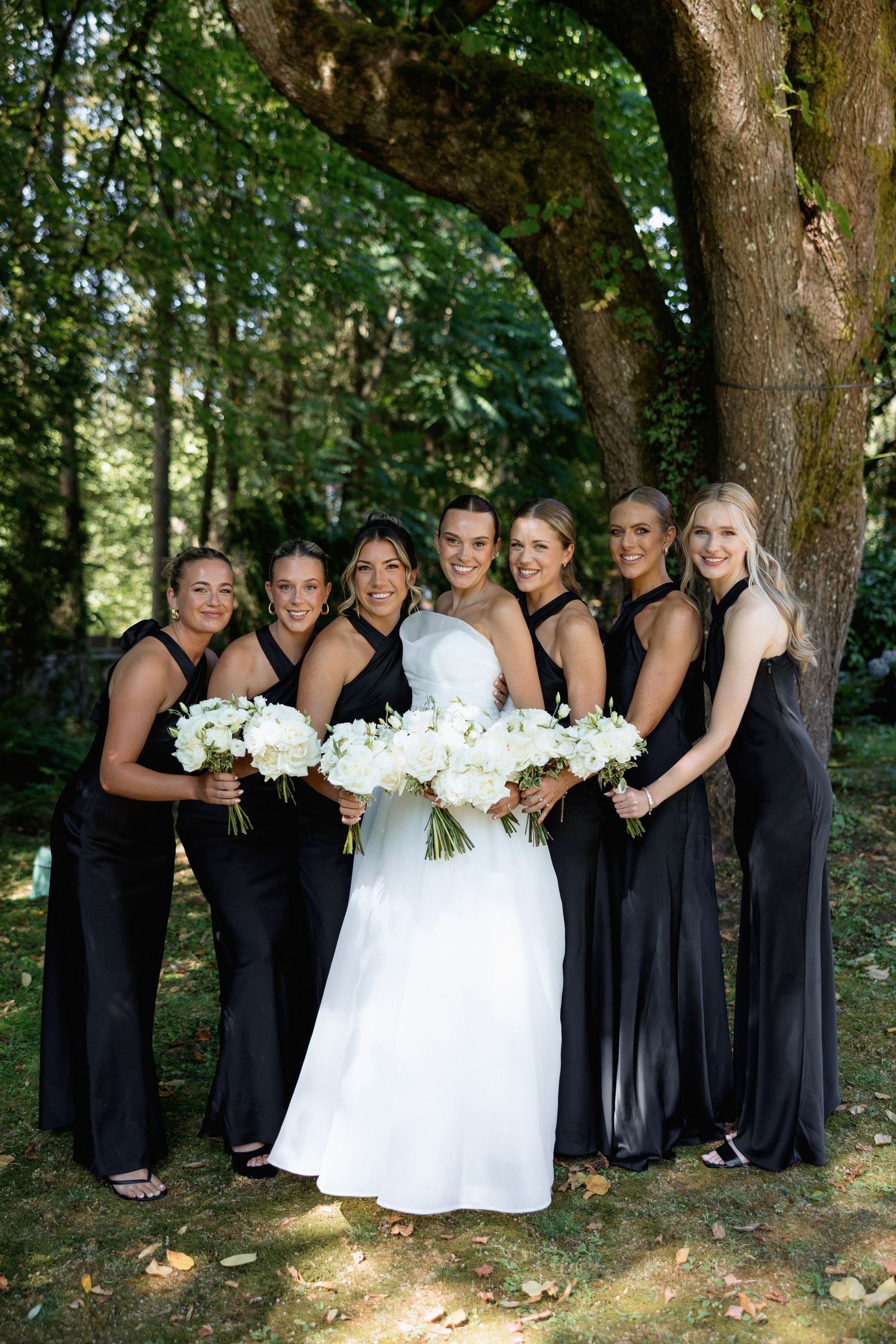 Bridal party in navy and white poses together under large trees.