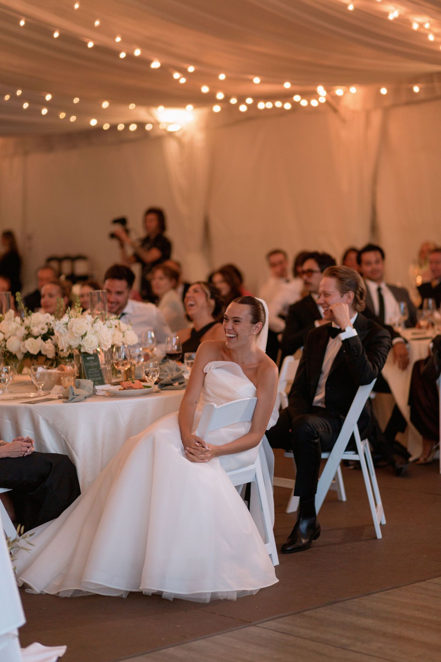 Wedding reception in a white tent decorated with string lights and elegant table settings.