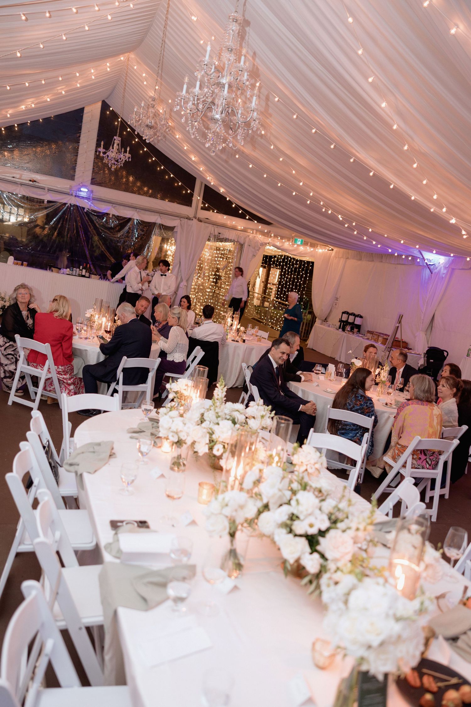 Elegant wedding reception setup with white tables, chairs and twinkling lights in a draped tent.