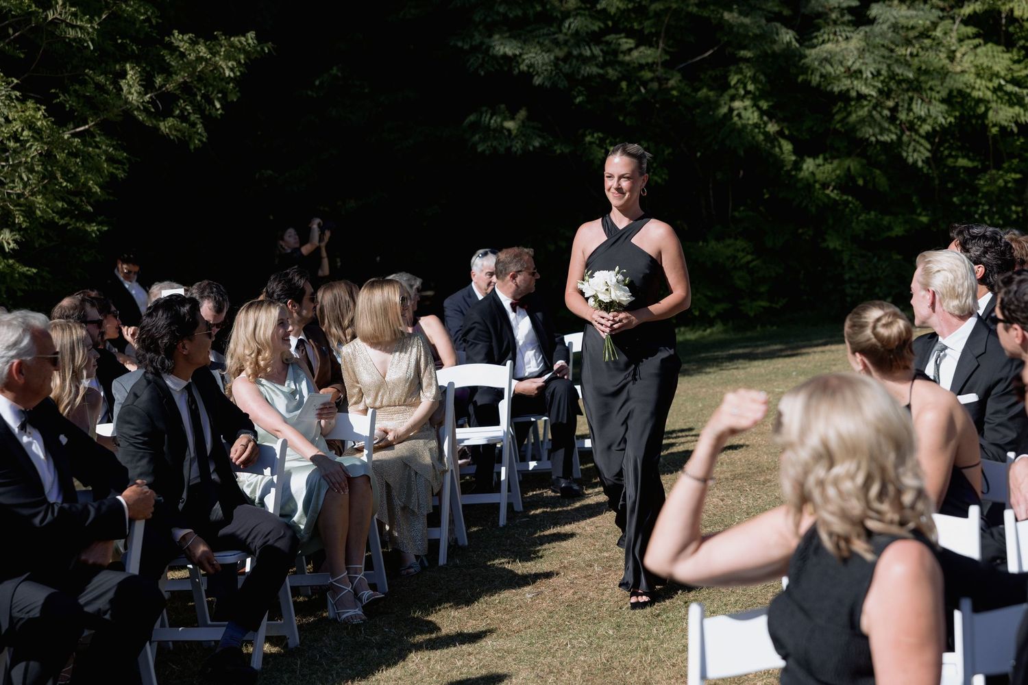 Wedding attendees stand in groups on a grassy area with white chairs arranged for an outdoor ceremony.