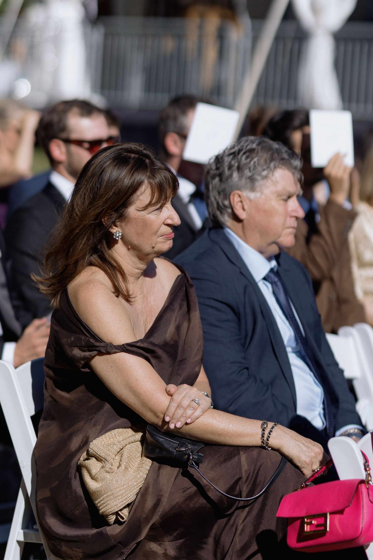 Guests seated at an outdoor wedding ceremony with a woman in brown dress and man in navy suit in focus.