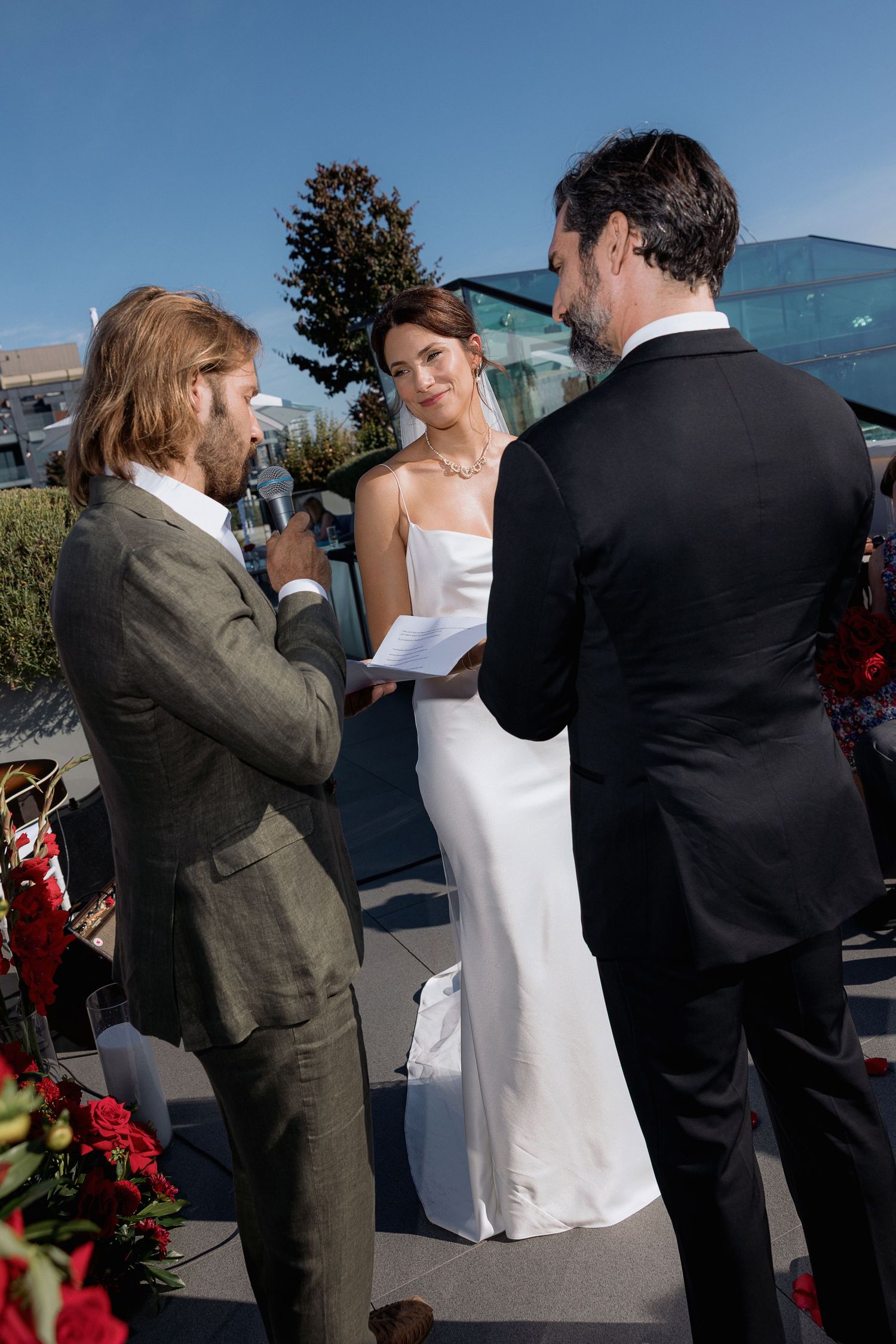 Wedding ceremony participants in formal attire gathered outdoors against a bright sky.