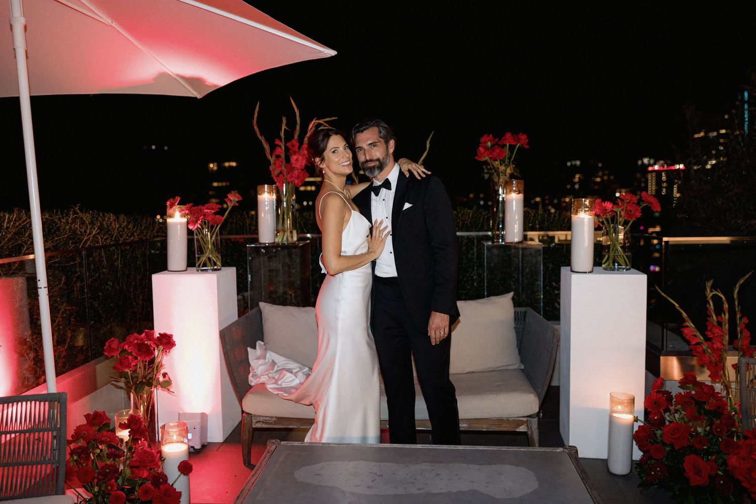 Couple posing on rooftop terrace with red floral arrangements and white decor in the night of Vancouver